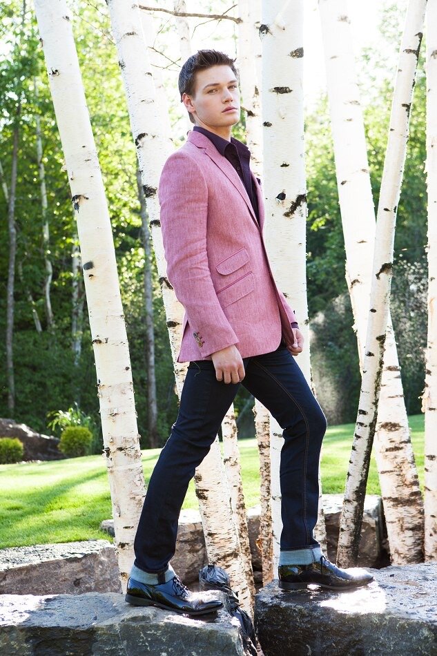 Young man standing on a rock among white birch trees in a forested park, wearing a pink blazer, black shirt, dark jeans, and dress shoes.