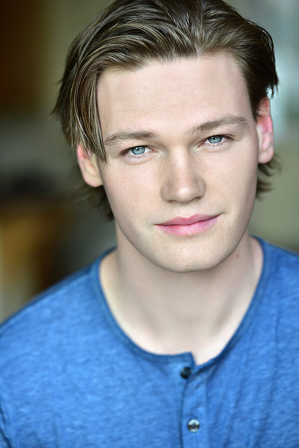 Close-up portrait of a young man with blue eyes, light skin, and brown hair, wearing a blue shirt, in a softly lit indoor setting.