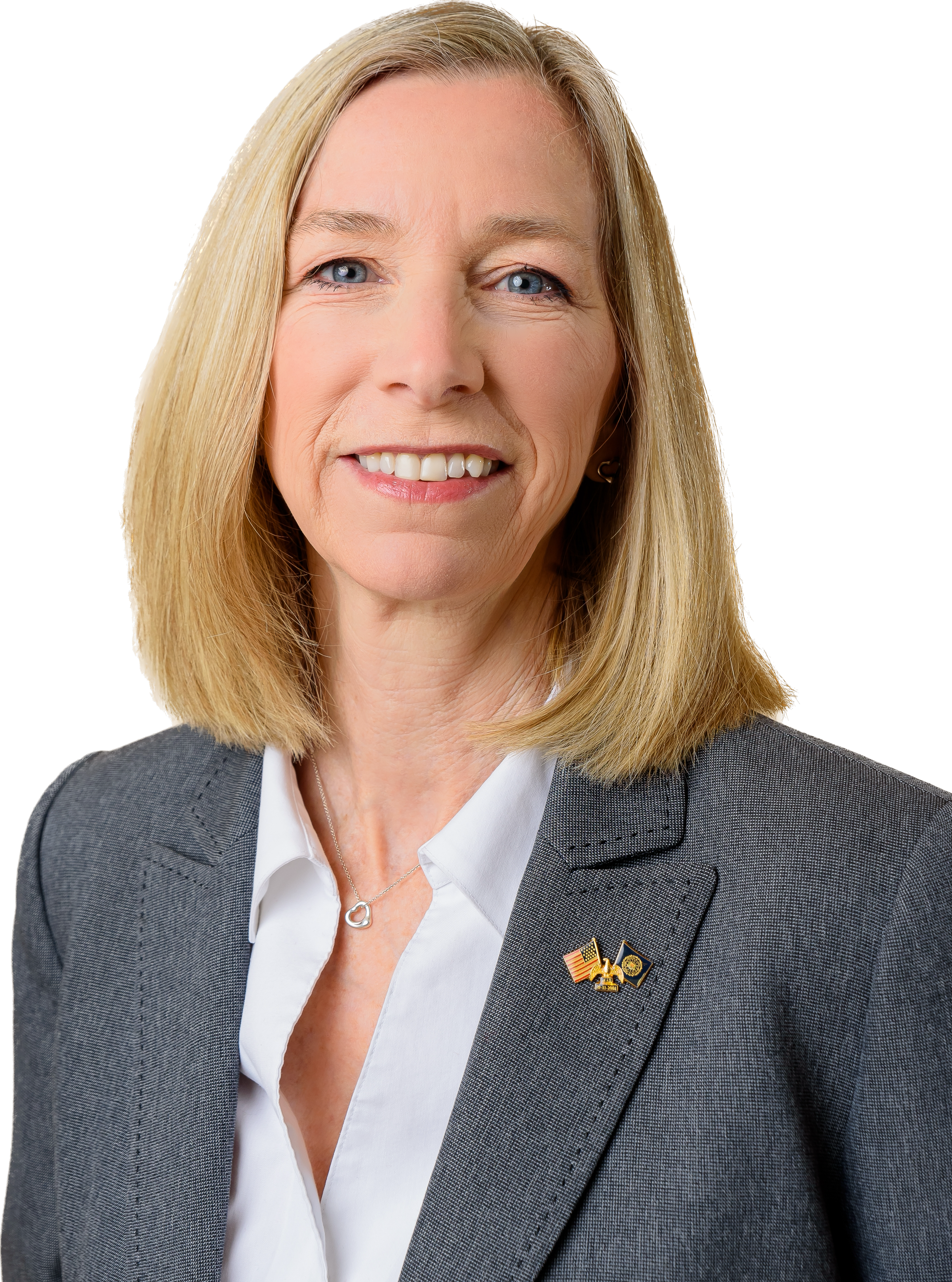 A middle-aged woman with shoulder-length blonde hair, wearing a gray blazer with a U.S. flag lapel pin, a white shirt, and a silver heart-shaped necklace, smiling confidently against a plain background.