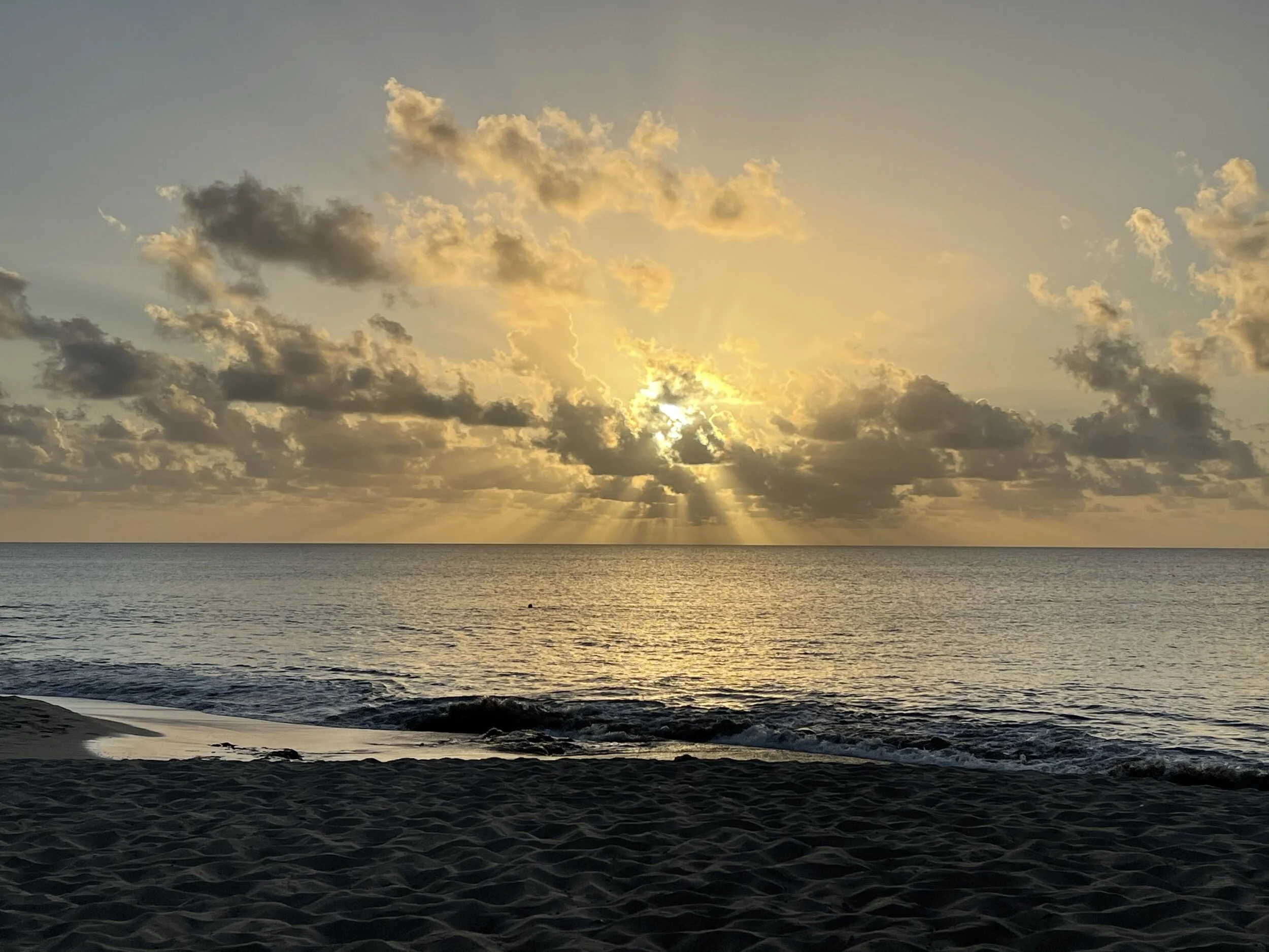 Sunset over ocean with sunbeams through clouds and sandy beach in foreground.