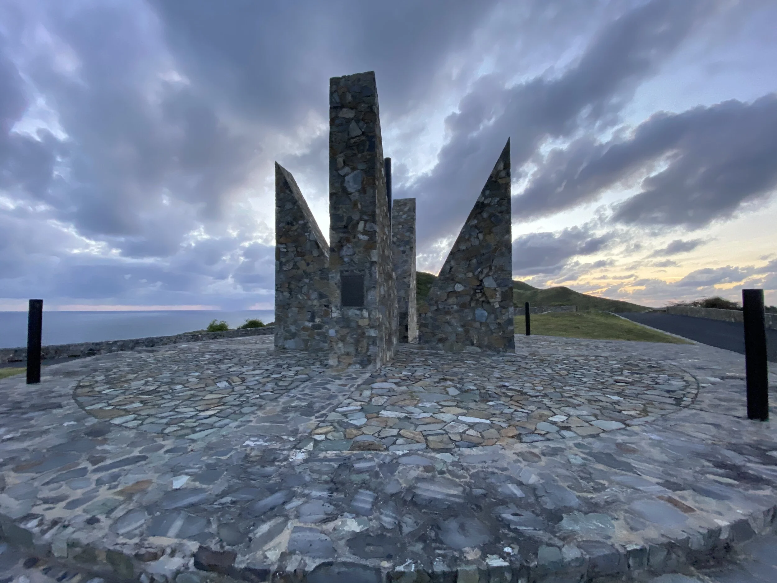 Stone monument with triangular structures on a circular stone platform, set against a cloudy sky and ocean backdrop.
