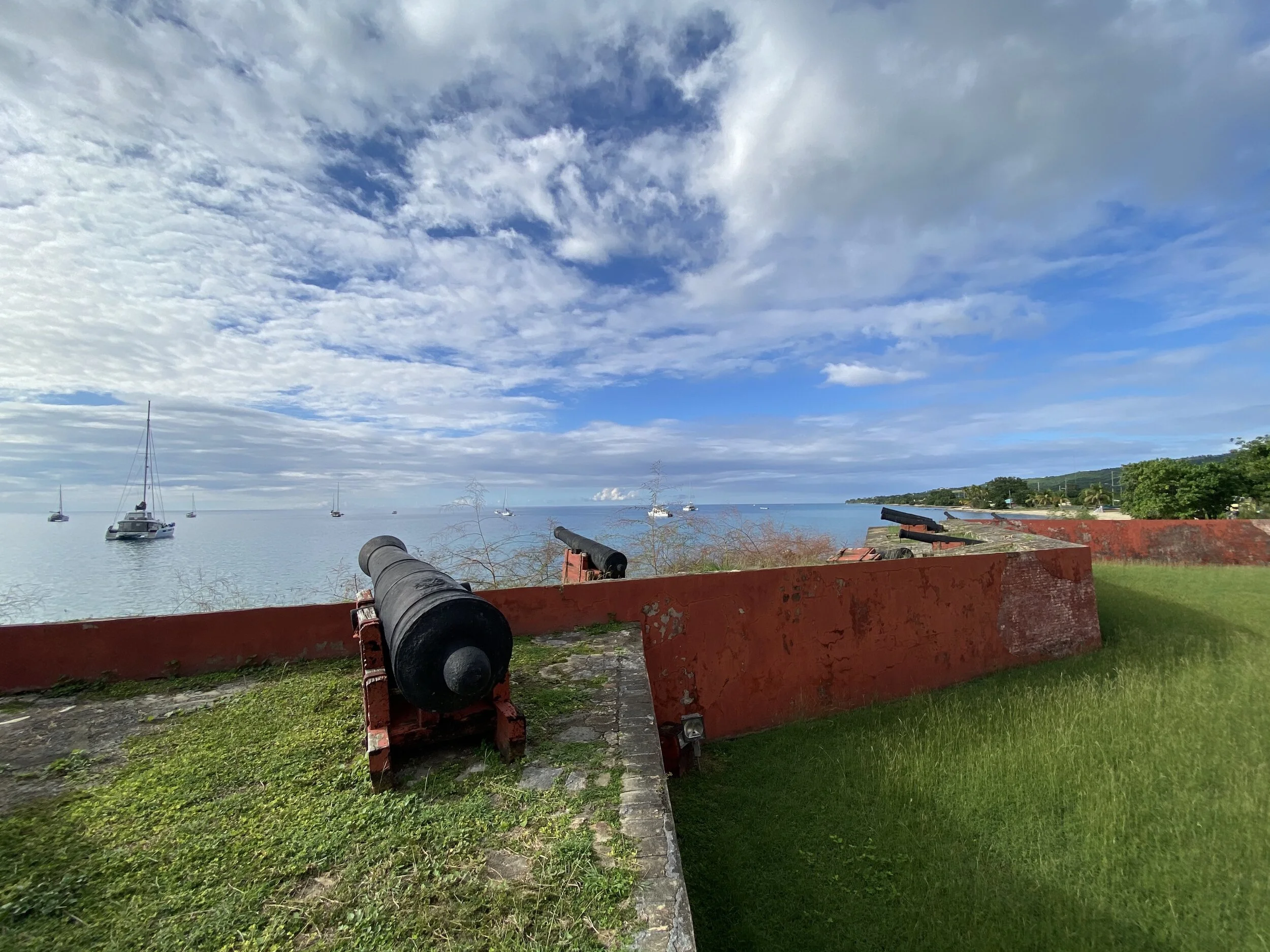 Fort with cannons overlooking a calm sea with sailboats in the distance, under a partly cloudy sky.