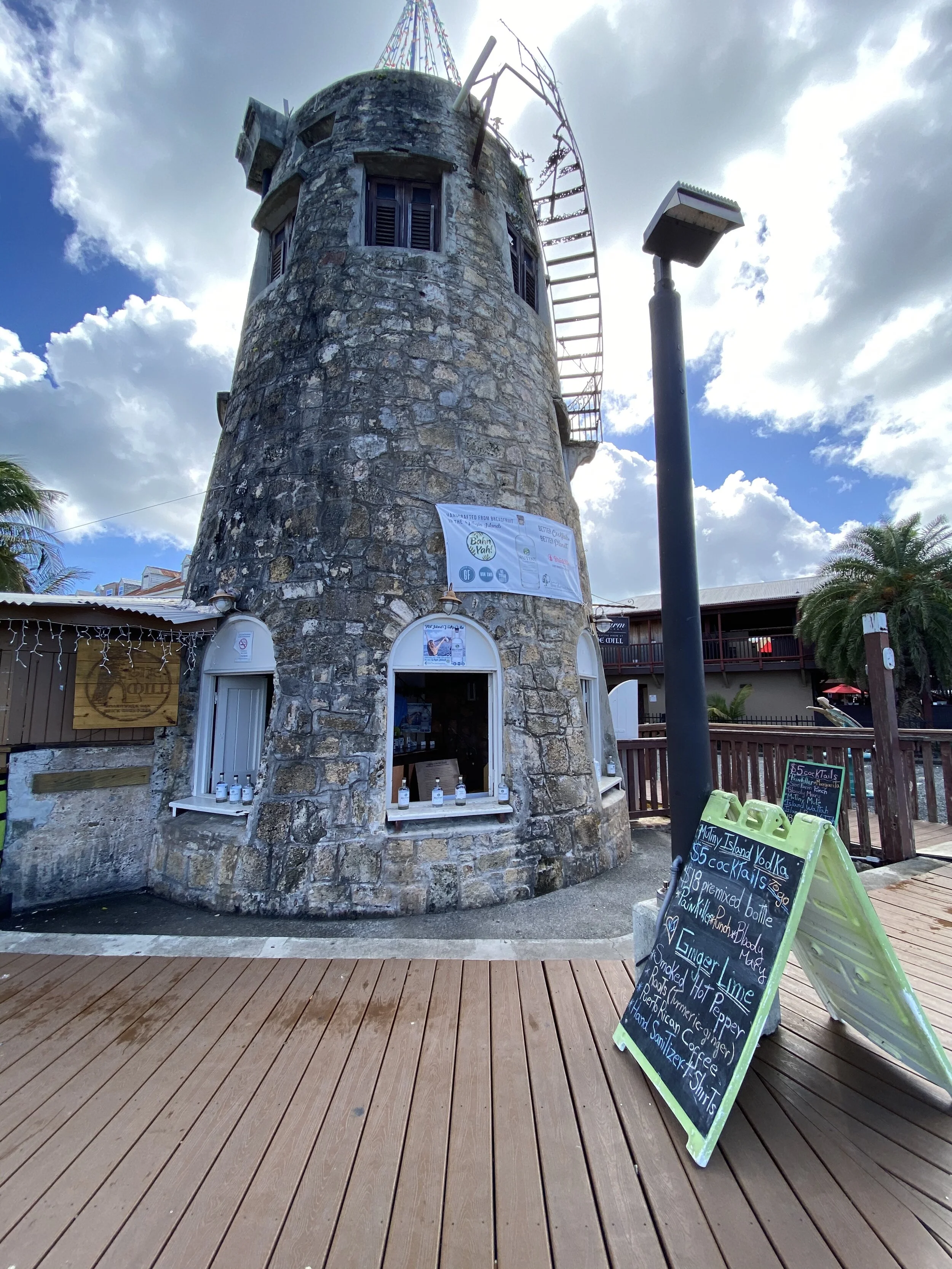 A stone windmill structure with a curved outer surface, featuring windows and a partially broken windmill blade at the top. In front of the building, there are signs with handwritten menu items and prices. The scene includes a wooden deck, light pole