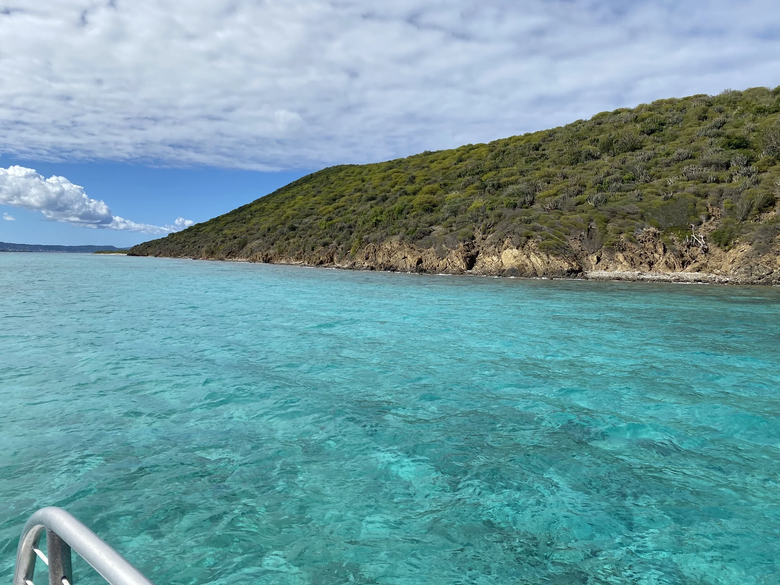 Clear turquoise water with a small mountain covered in green vegetation and a partly cloudy sky in the background.
