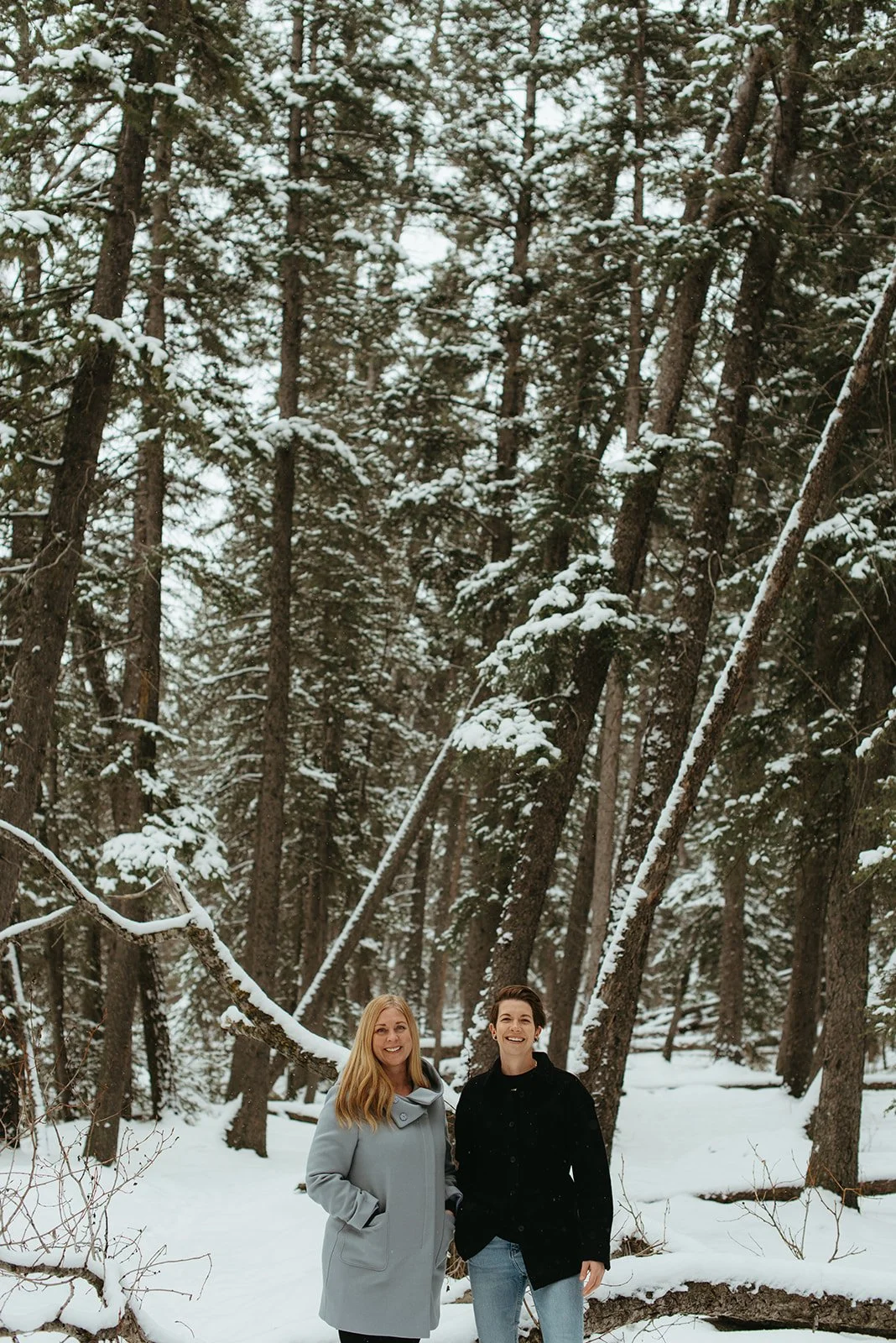 Two women standing in a snow-covered forest with tall pine trees in the background.