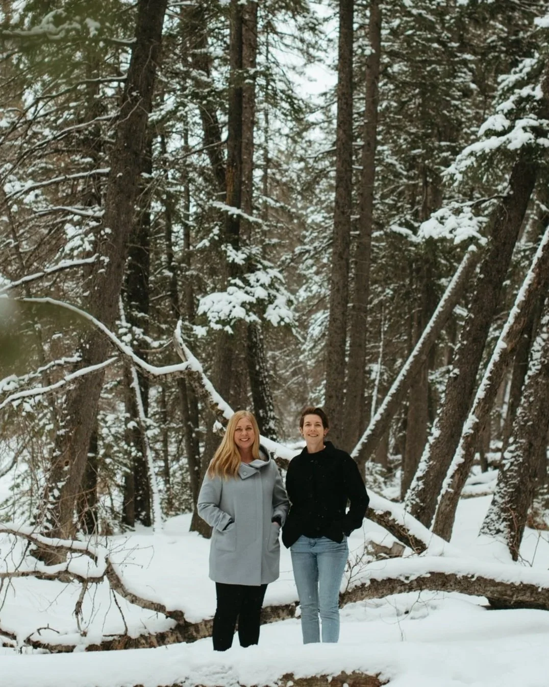 Two women standing in a snowy forest with tall trees and snow-covered branches, smiling and posing for the photo.
