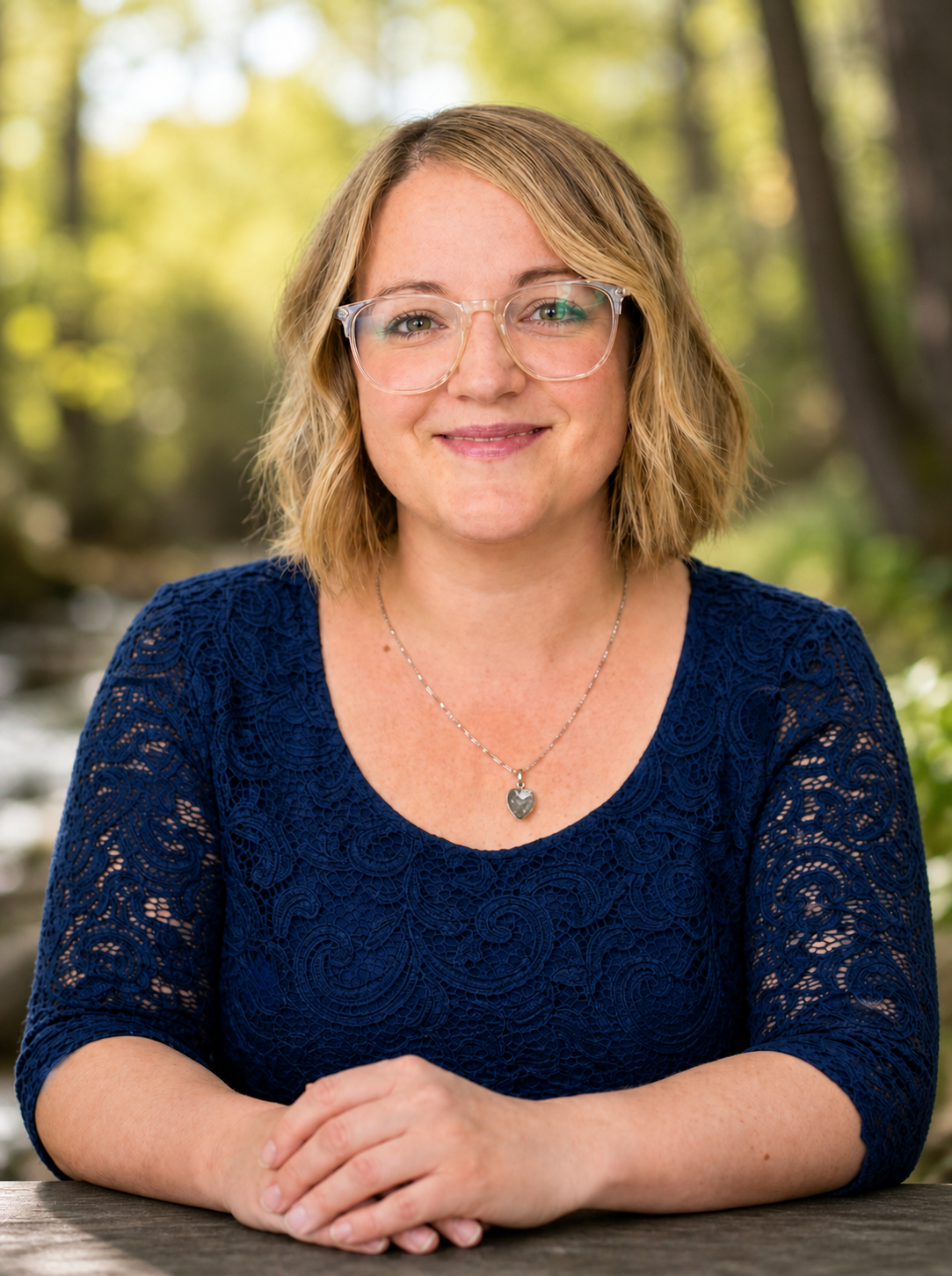 A woman with shoulder-length blonde hair, wearing glasses and a blue lace top, sitting outdoors at a wooden table with green trees in the background.