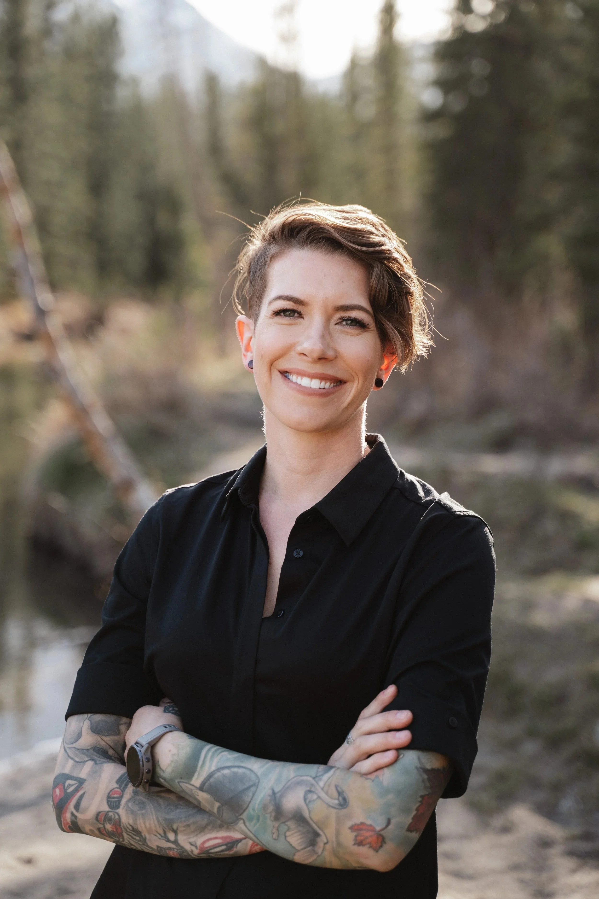 A woman with short, styled hair, smiling with teeth, wearing a black shirt and sporting tattoos on both arms, standing outdoors near a body of water with trees in the background.