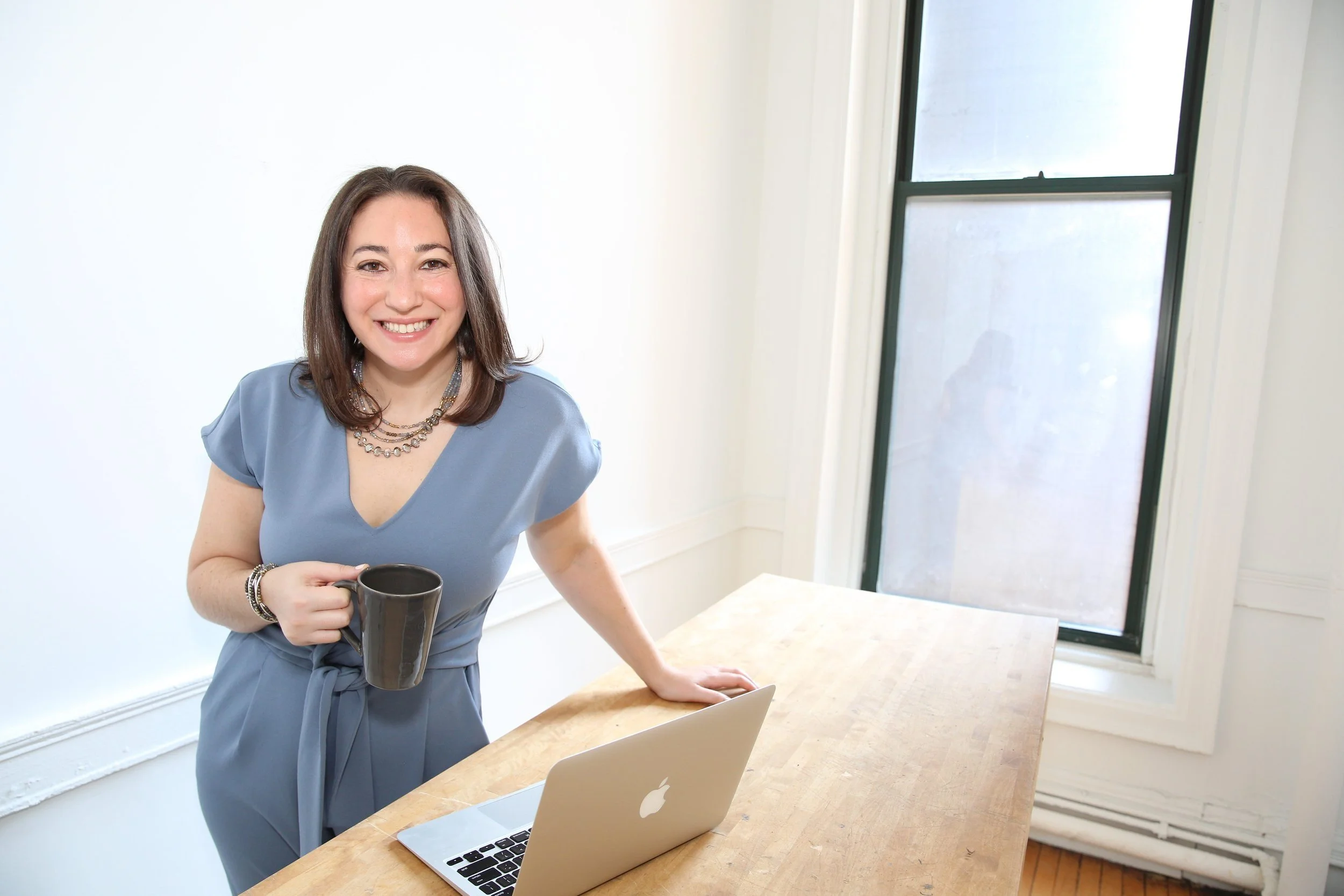 A woman with brown hair smiling, wearing a light blue dress and jewelry, holding a coffee mug, standing in a bright room with a wooden table, an open Apple laptop, and a large window.