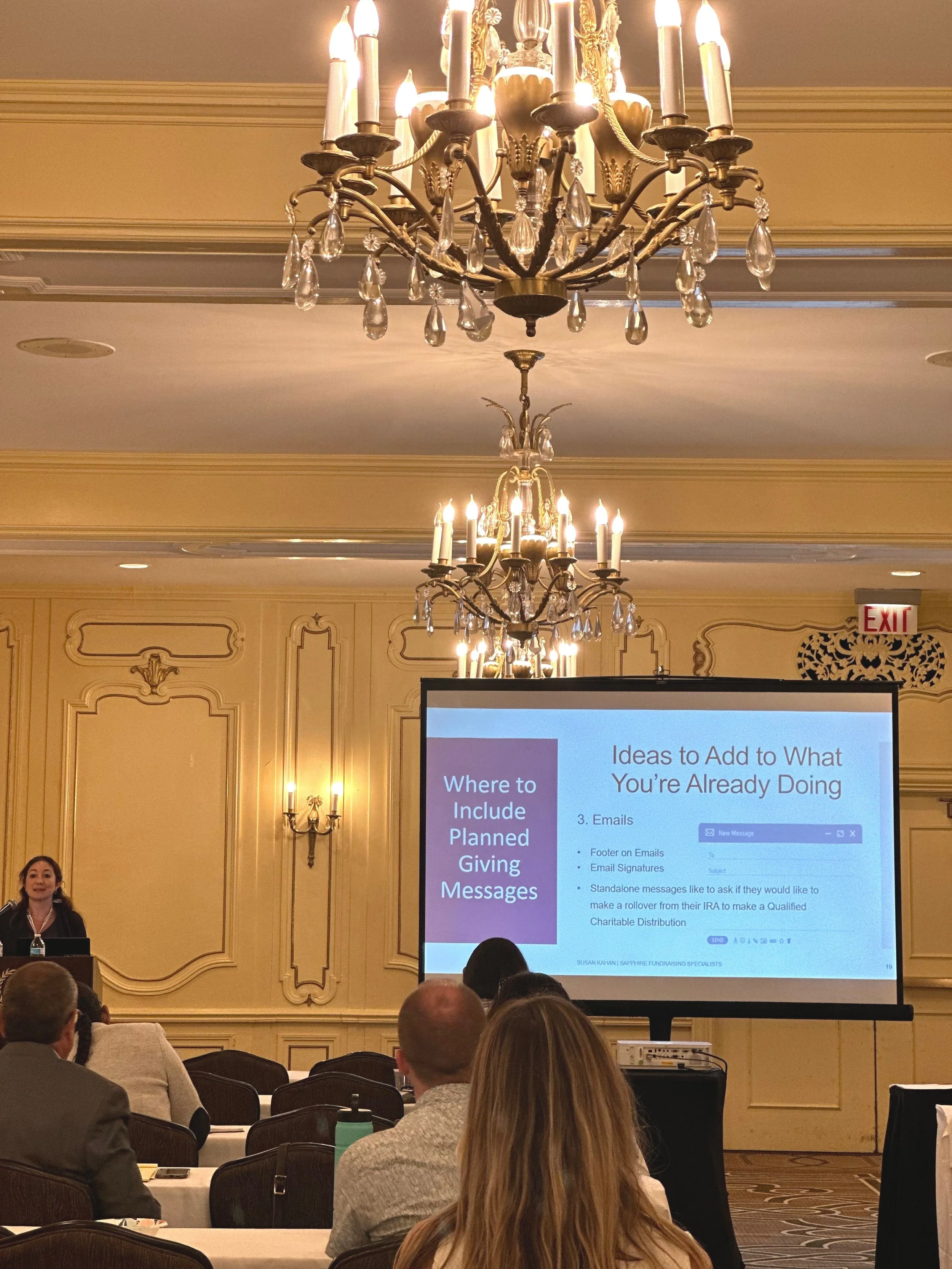 Conference room with a presentation on planned giving. The slide includes text about adding ideas to emails for planned giving messages. The room is elegantly decorated with chandeliers and attendees are seated, focused on the presentation.