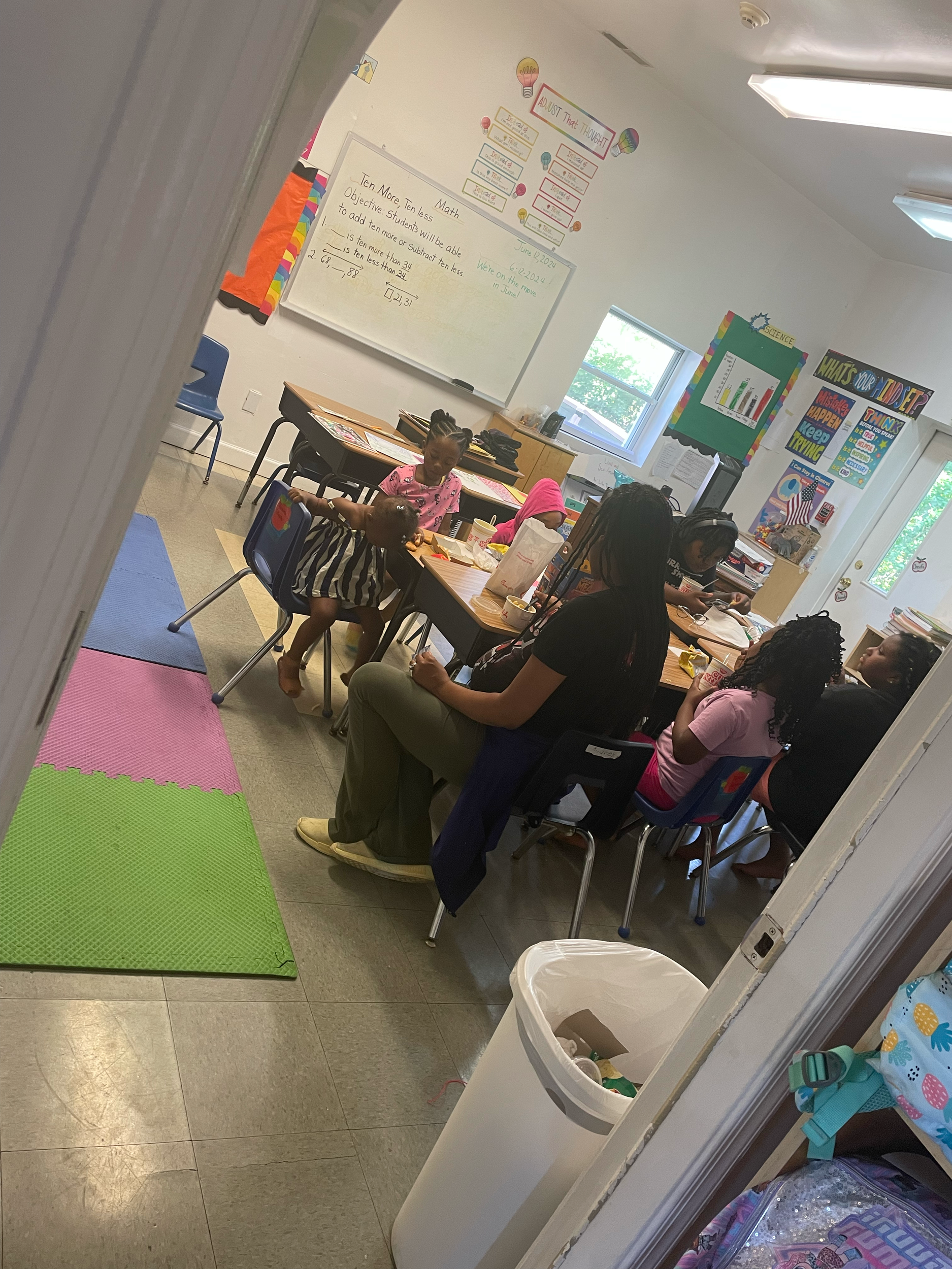 A classroom viewed from a doorway with students and a teacher seated at tables, studying or working on assignments. The room has bright lighting, educational posters, and a whiteboard on the wall.