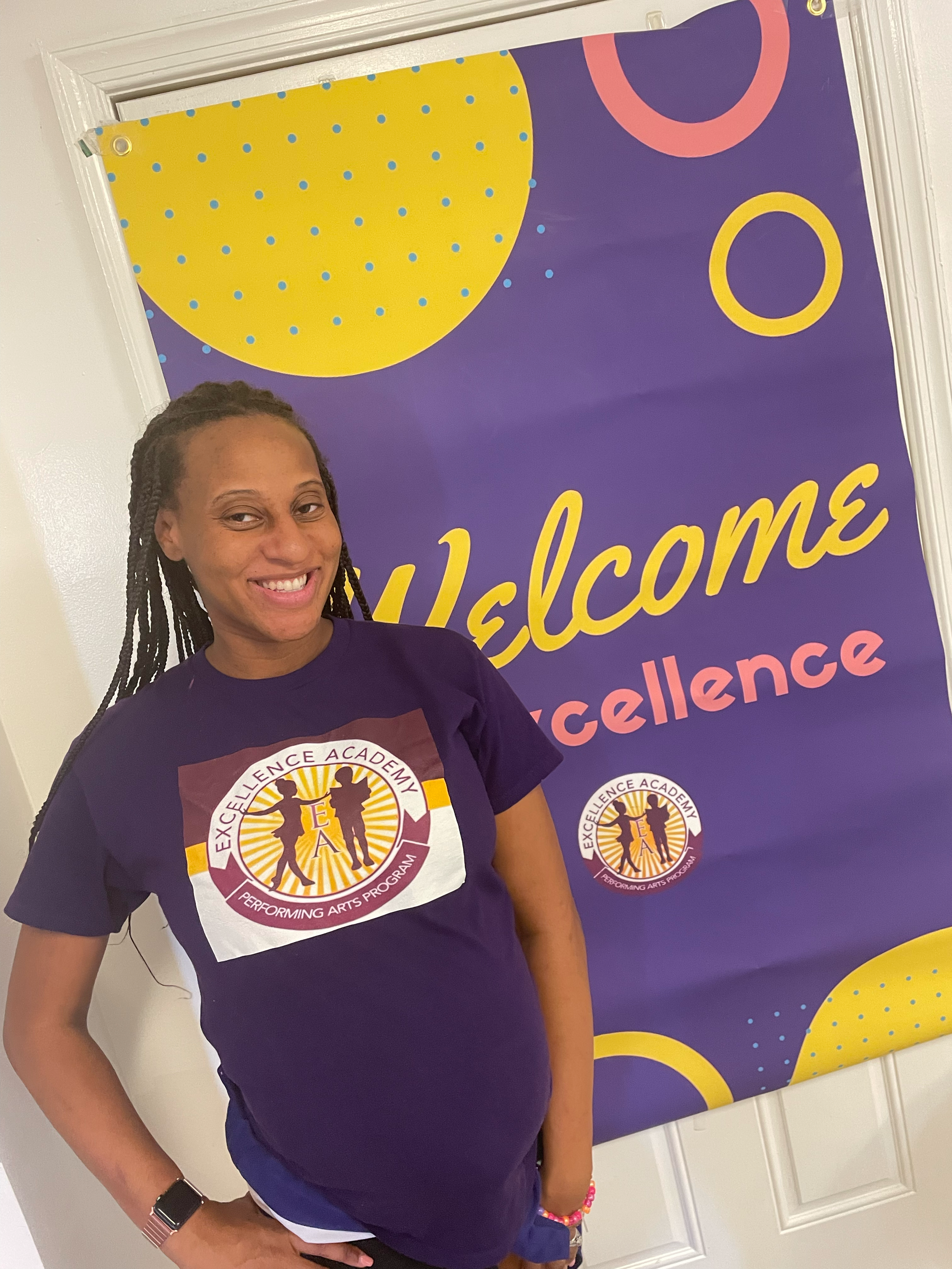 A smiling woman with long braided hair standing in front of a welcome banner for 'Excellence.' She is wearing a purple t-shirt with the Excellence Academy logo and a smartwatch on her left wrist.