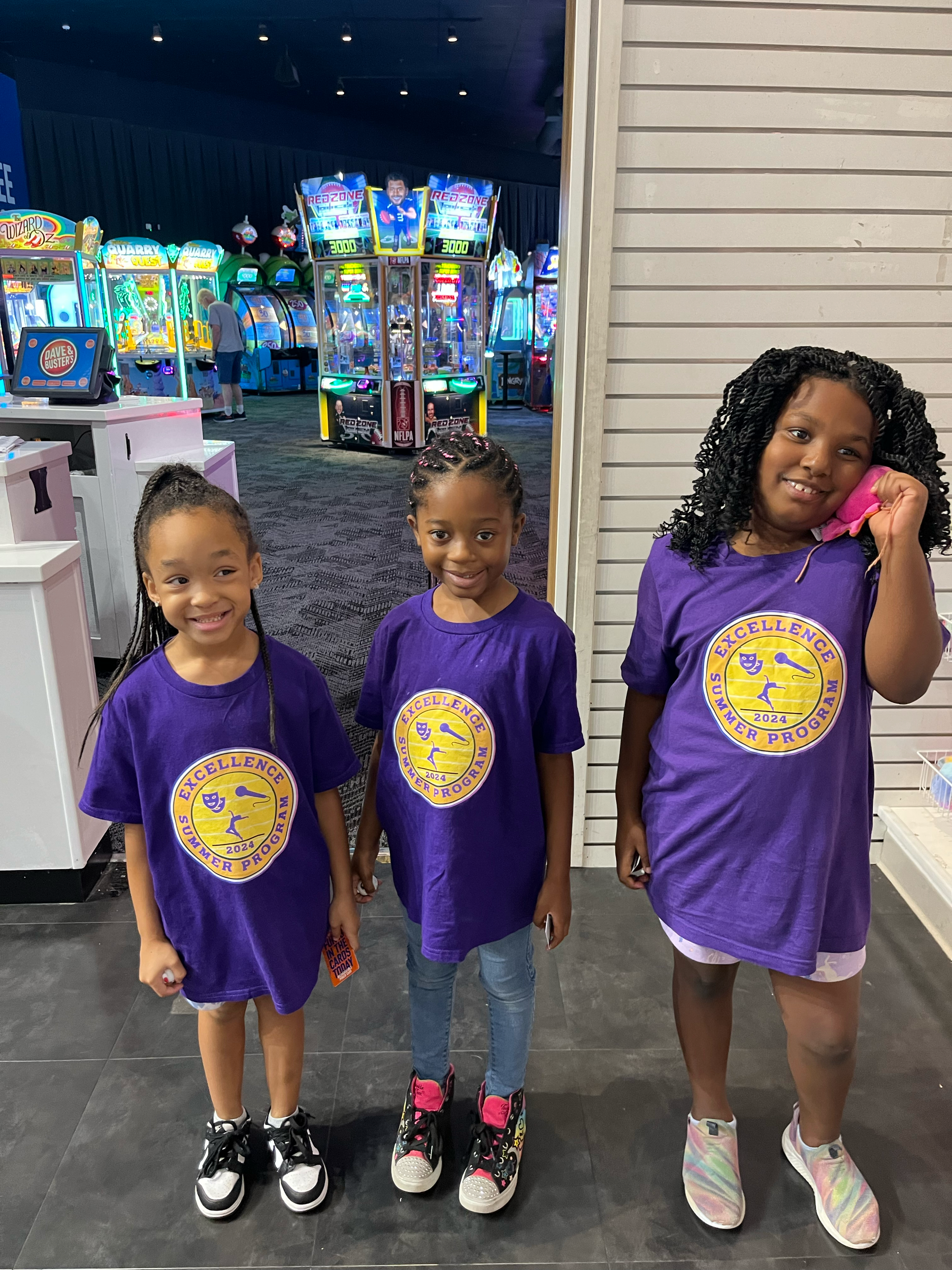 Three young girls in purple shirts with 'Excellence Summer Program 2024' logo, standing in arcade with game machines and attractions in the background, one girl talking on a pink phone.