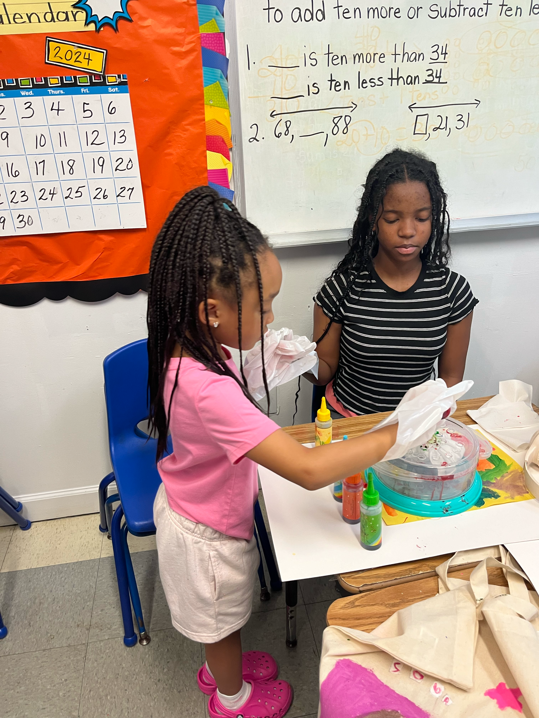 Two young girls are engaging in an activity at a classroom table with bottles of colored paint, tissue paper, and a spinning set with beads, possibly for a craft project. One girl, in a pink shirt and shorts, is handing tissue paper to the other girl