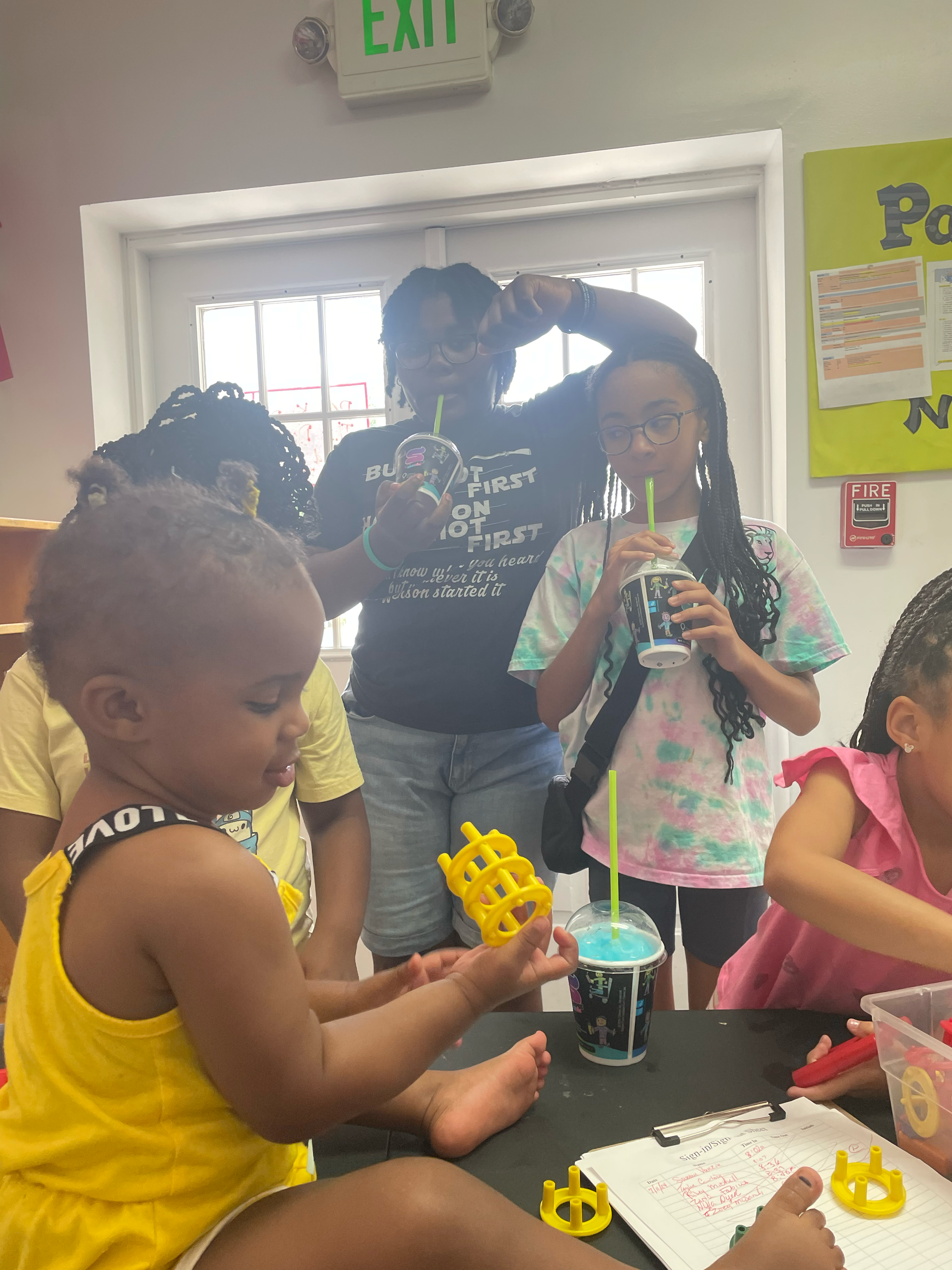 Children and a woman in a room with a window, wearing colorful clothes and glasses, enjoying drinks with straws. A child in yellow is playing with yellow plastic toys while others are reaching for items on the table. The room has a green exit sign an