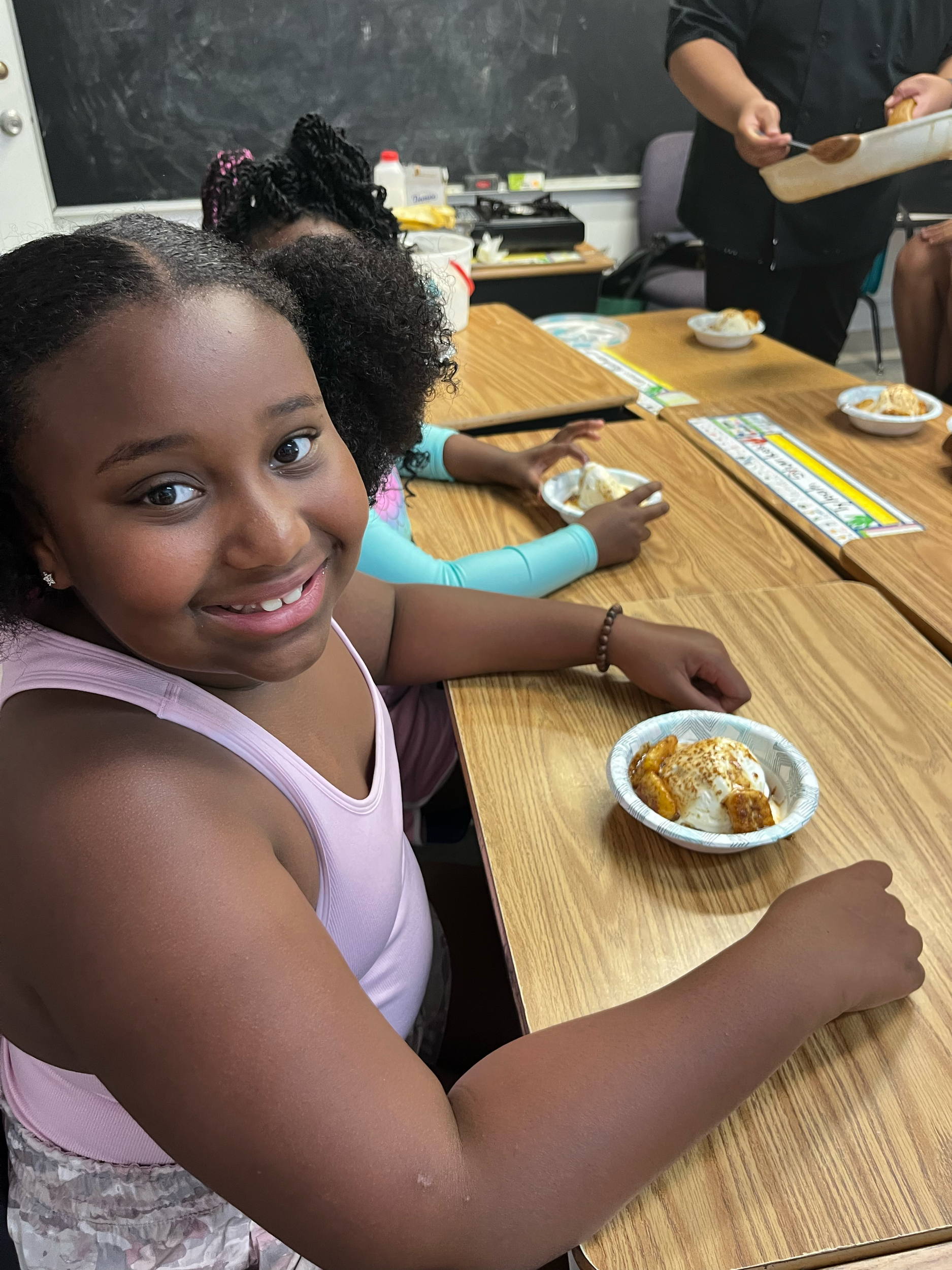 A smiling girl with curly hair sitting at a table with a plate of food in front of her, with other children and adults in the background during a meal or snack event in a classroom.