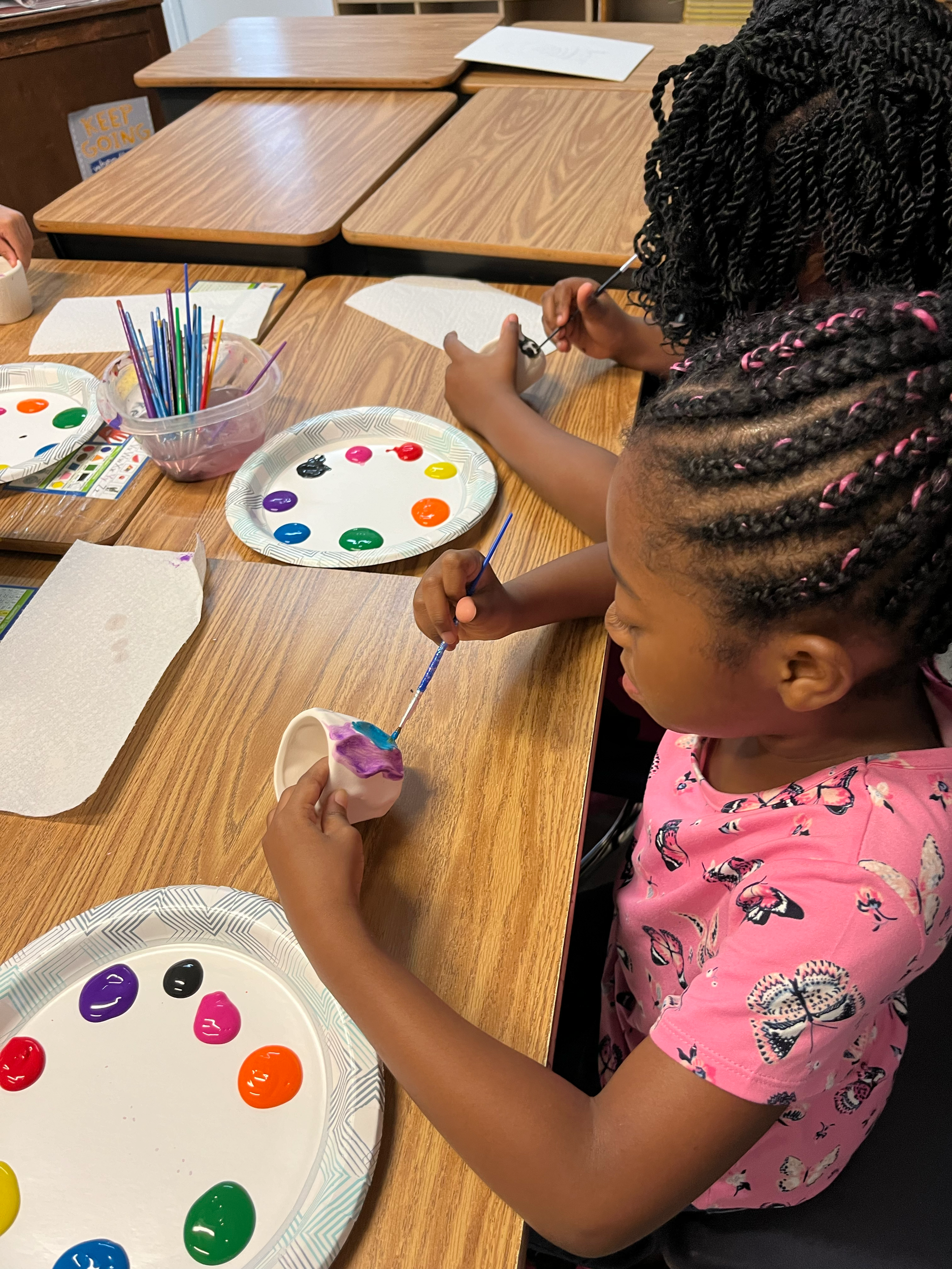 Two children painting ceramic cups with colorful paint at a wooden table.