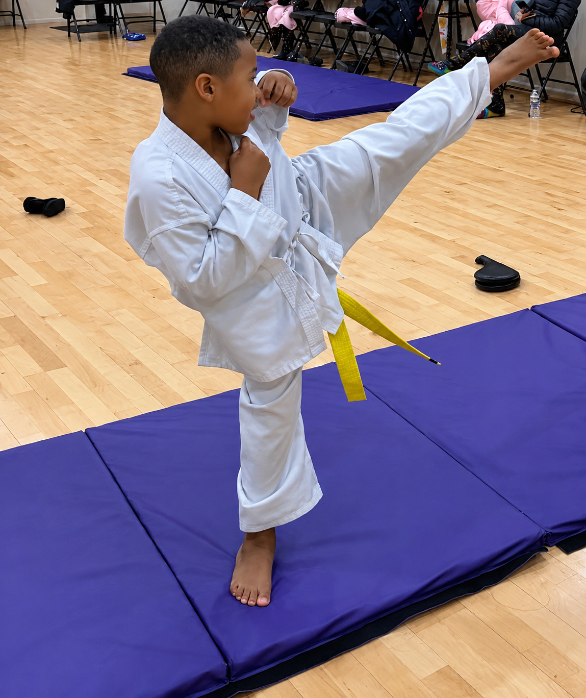 A young boy wearing a white martial arts uniform performing a high kick in a classroom.