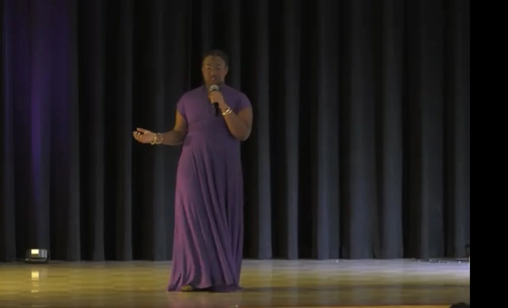 A woman in a red gown holding a microphone and raising her hand in a church or auditorium setting with wooden pews and white walls.