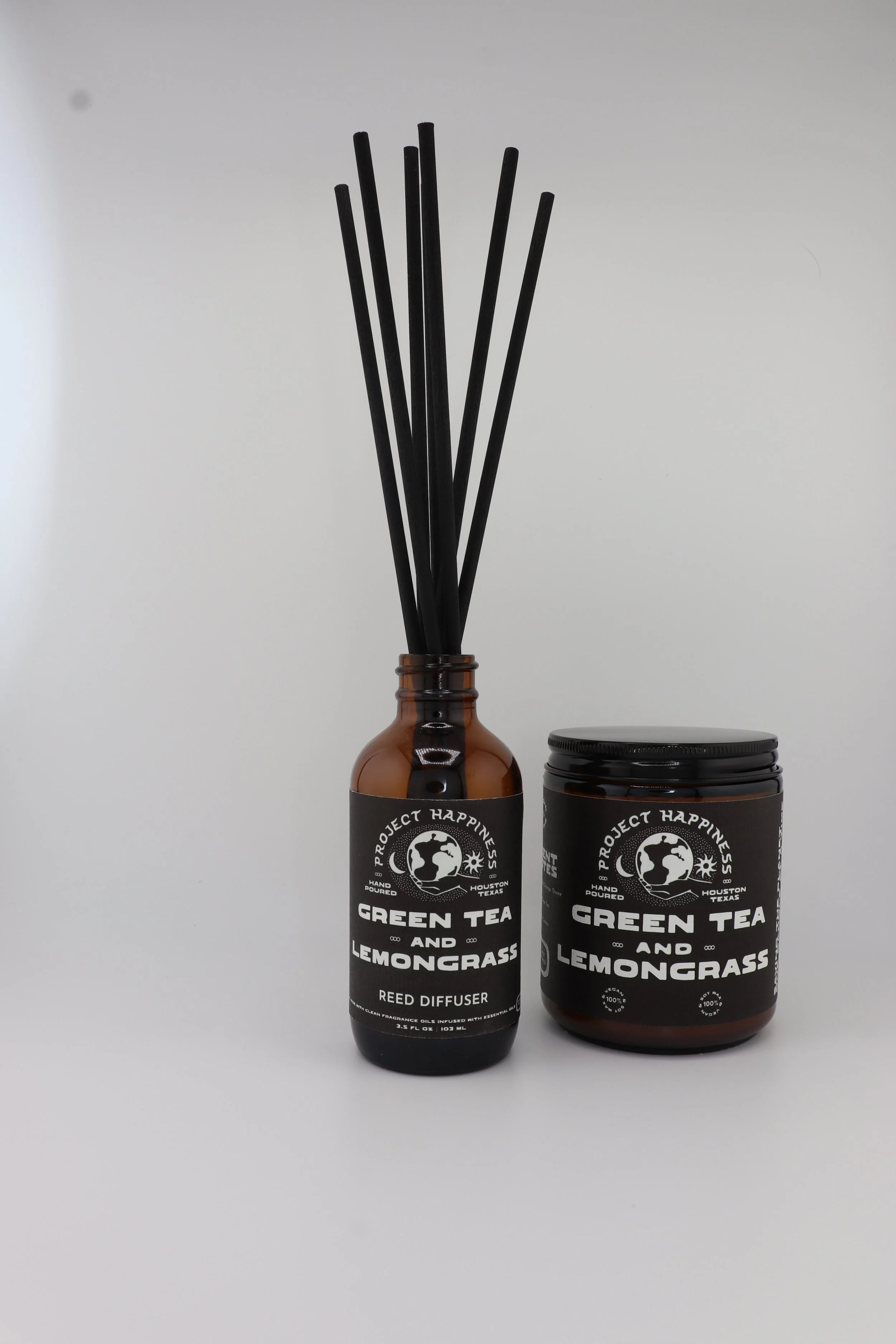 Green Tea & Lemongrass in amber glass reed diffuser with black label and black reed sticks, placed next to a matching amber jar candle with black label, on a clean white background.