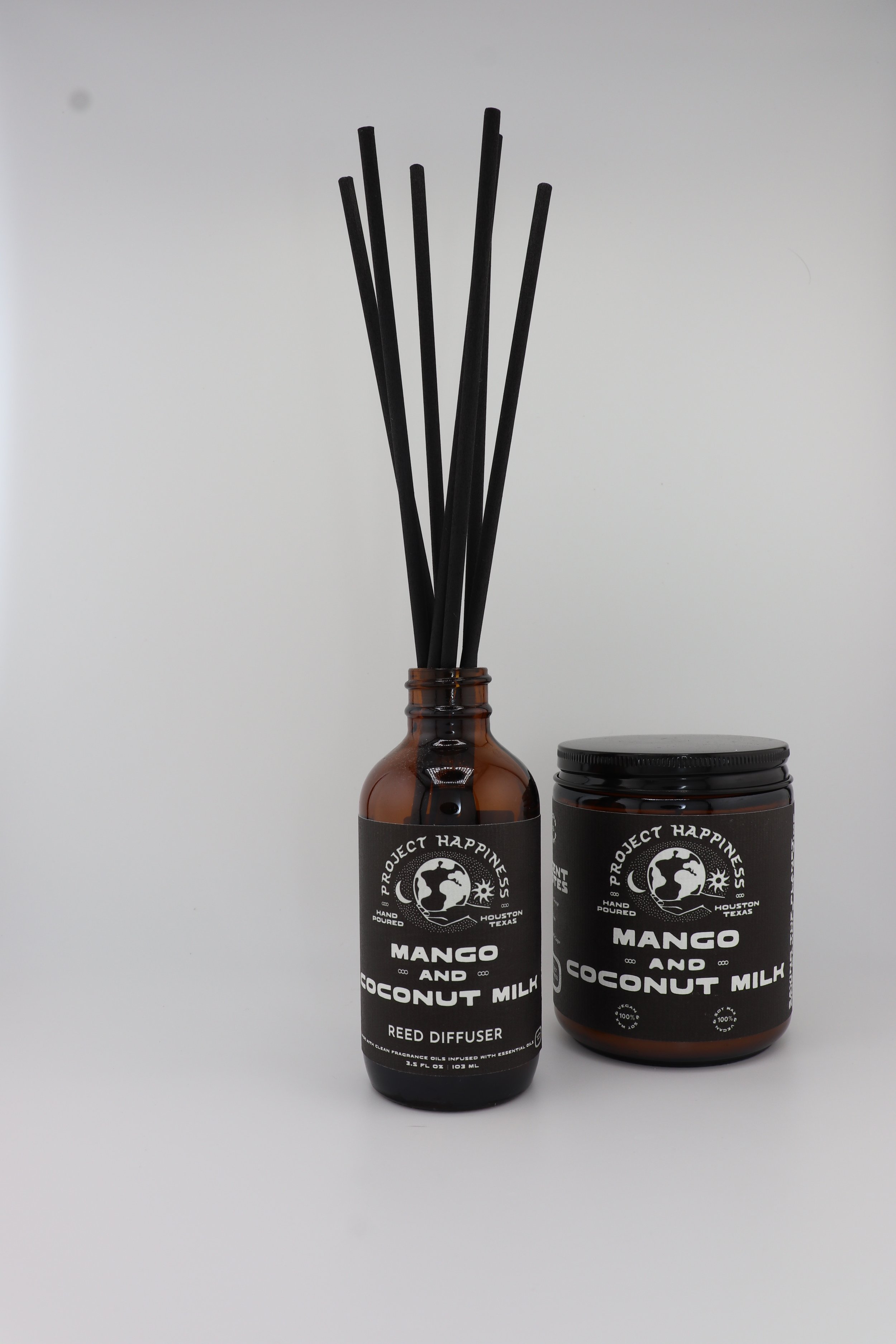 Mango & Coconut Milk in amber glass reed diffuser with black label and black reed sticks, paired with a matching amber jar candle with black label, on a white background.