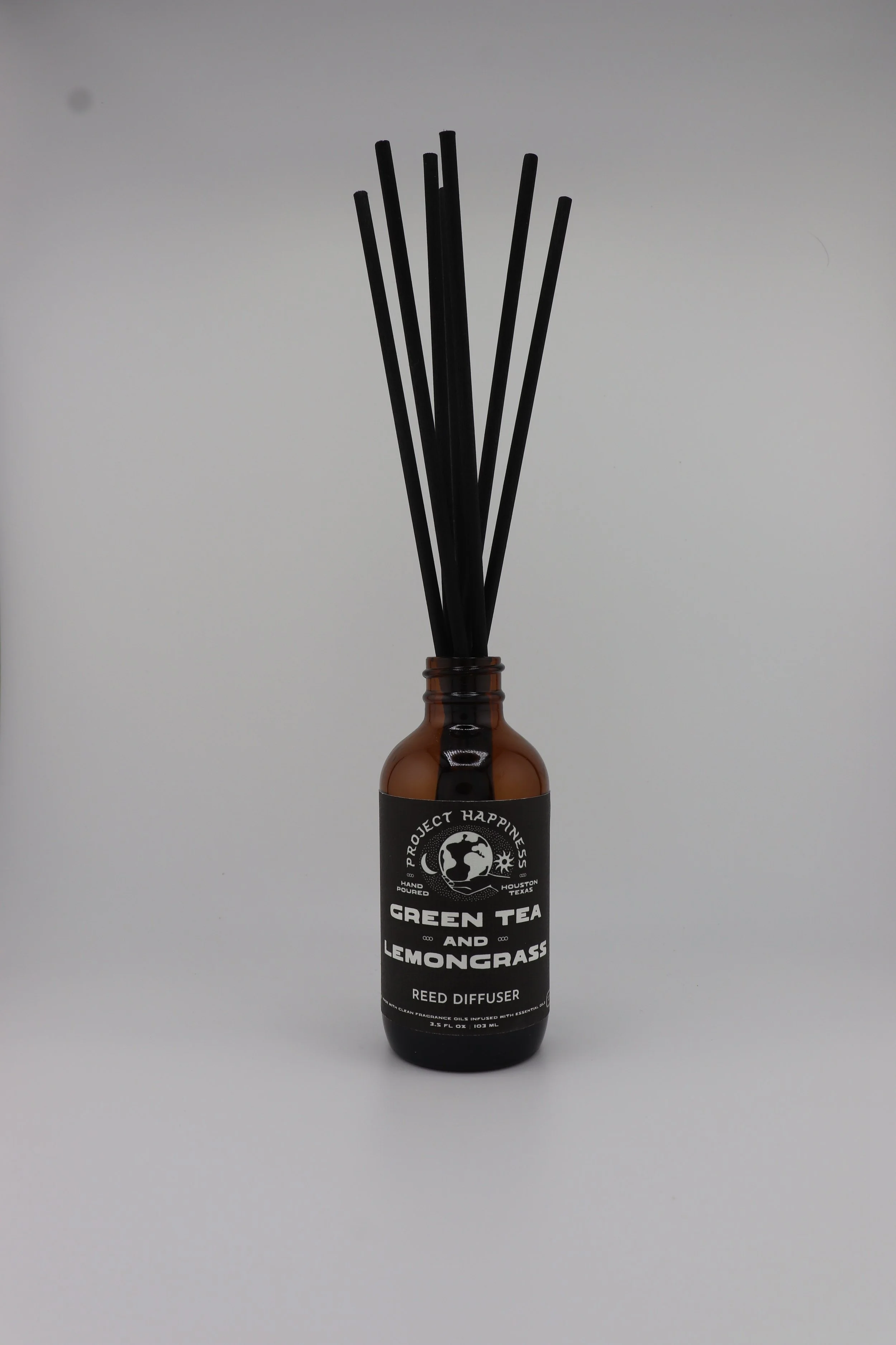 Amber glass reed diffuser with black label and black reed sticks on a white background, scented with green tea and lemongrass.