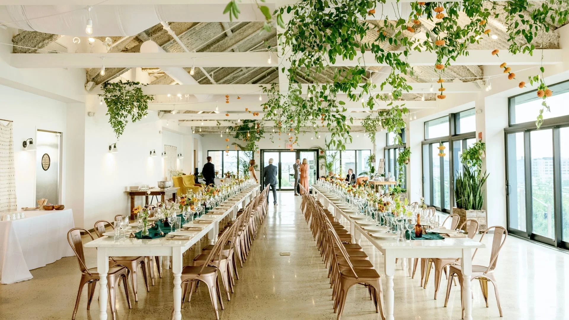 Long banquet tables with floral centerpieces and glassware in a brightly lit modern event space with greenery hanging from the ceiling.
