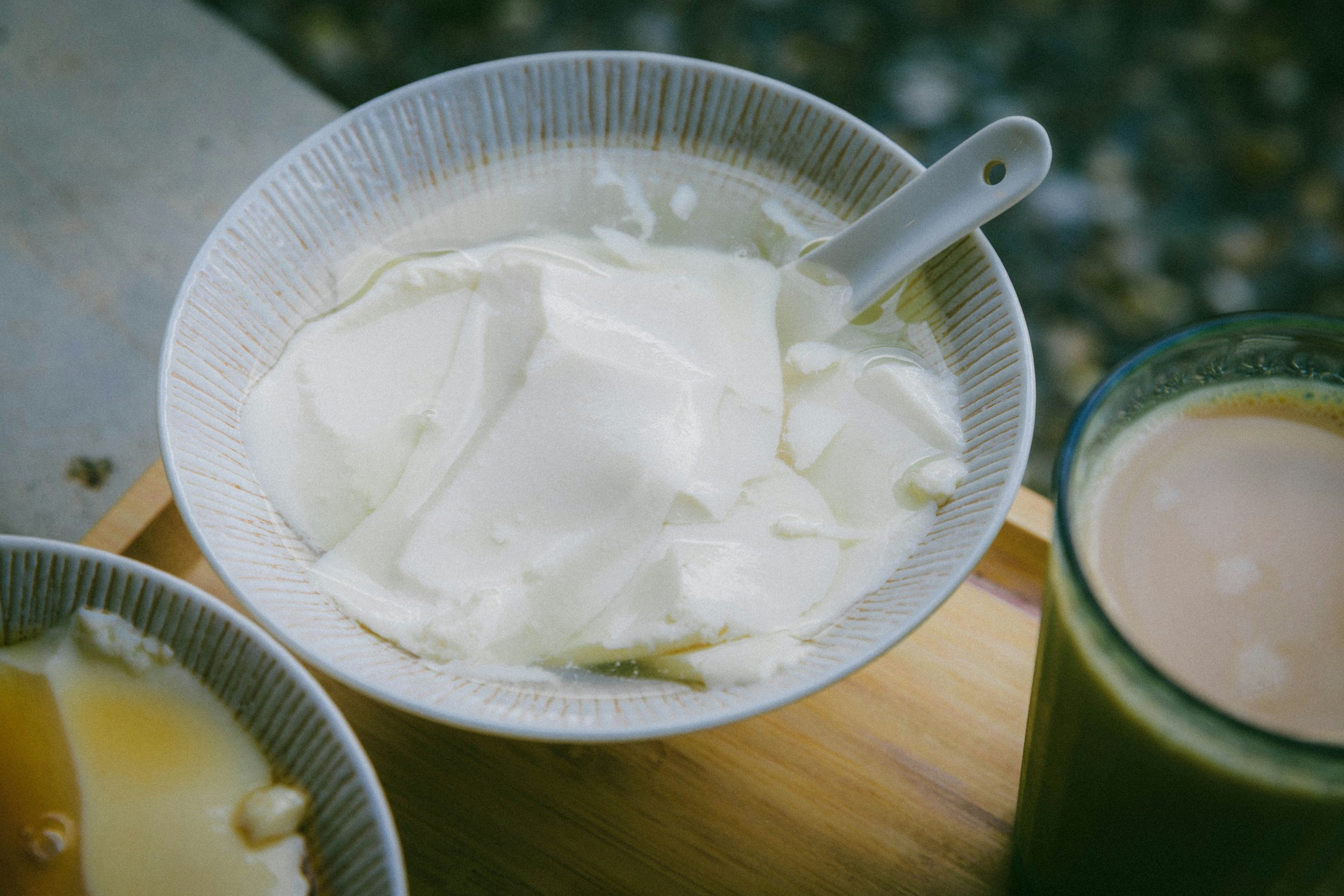 A bowl of yogurt with slices of pieces in a white bowl with a handle, a glass of beige-colored smoothie or juice, and another bowl of a similar yellowish pudding or dish on a wooden surface.