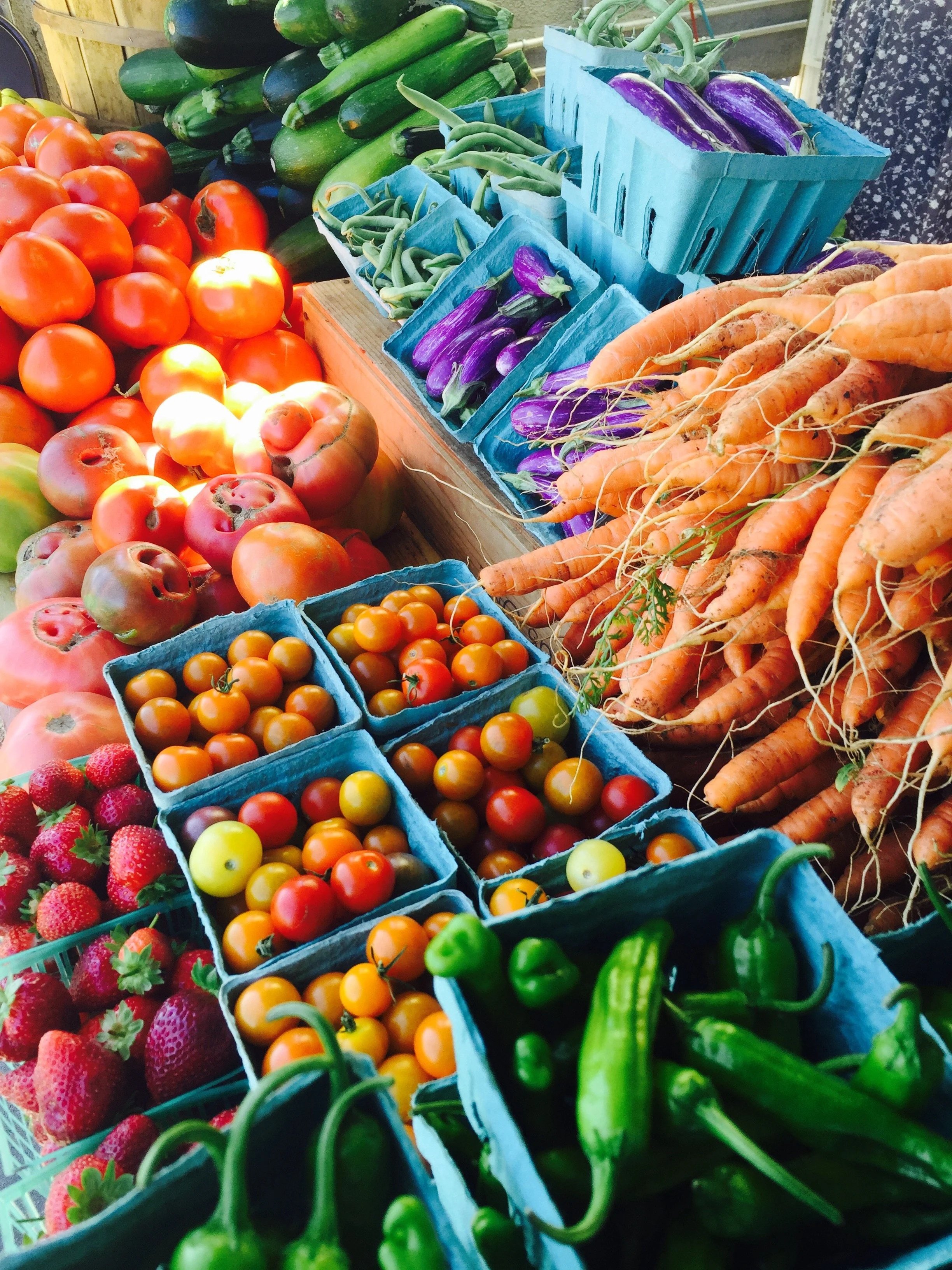 Fresh vegetables and fruits at a market stand, including tomatoes, strawberries, zucchinis, eggplants, carrots, and peppers arranged in baskets and boxes.