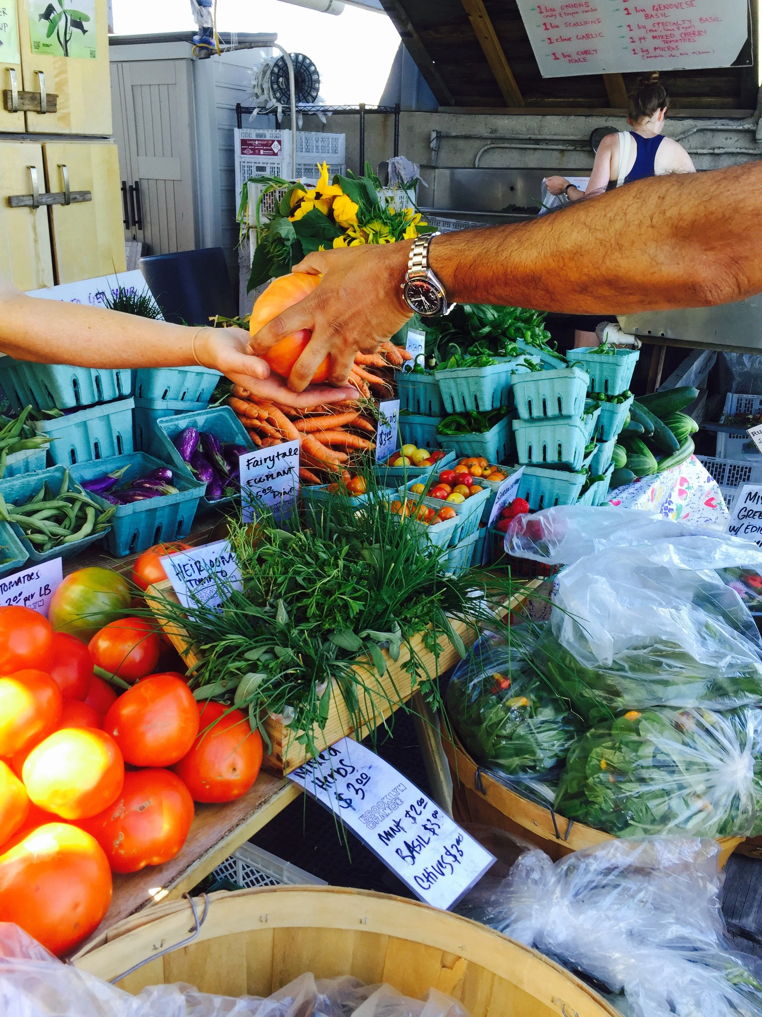 Two people exchanging an orange tomato at a farmer's market stand filled with fresh vegetables including tomatoes, carrots, eggplants, and leafy greens.