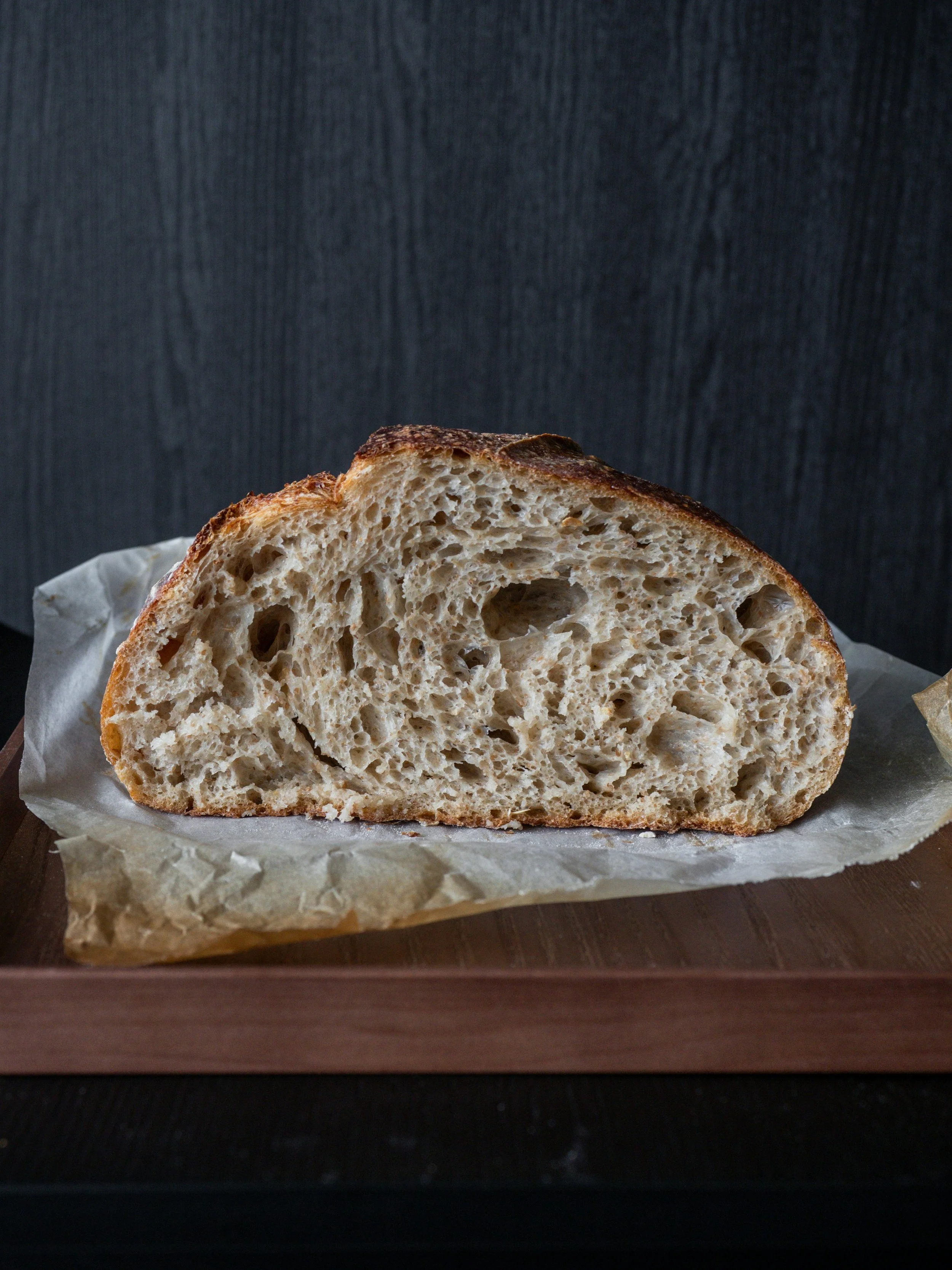 Close-up of a loaf of sourdough bread with a crunchy crust, sliced in half, on parchment paper on a wooden tray.