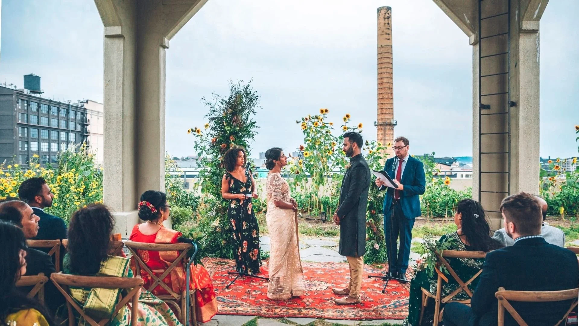 A diverse group of people participating in a wedding ceremony with the couple exchanging vows on a rooftop garden under an open pavilion, with guests seated on wooden chairs watching, surrounded by sunflowers and greenery.