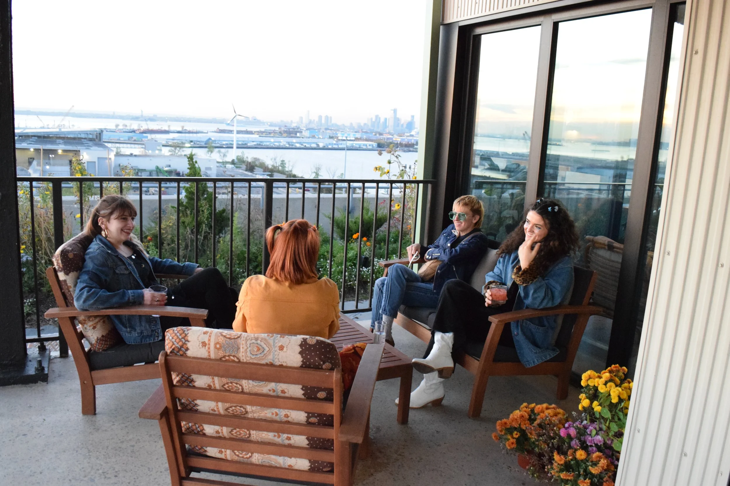 Four women sitting on a balcony with a cityscape and water in the background, enjoying drinks and conversation.