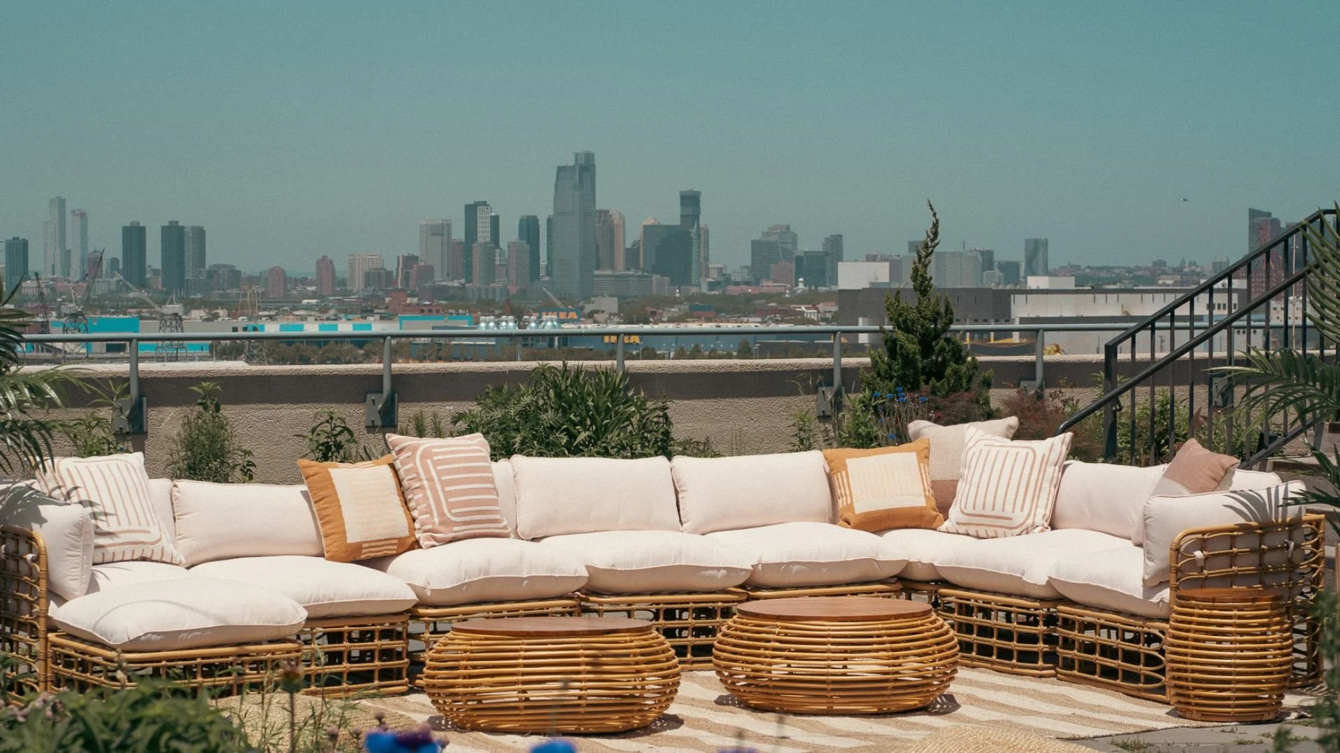 Outdoor rooftop seating area with white cushioned sectional sofa and rattan coffee tables, overlooking a city skyline with tall buildings.
