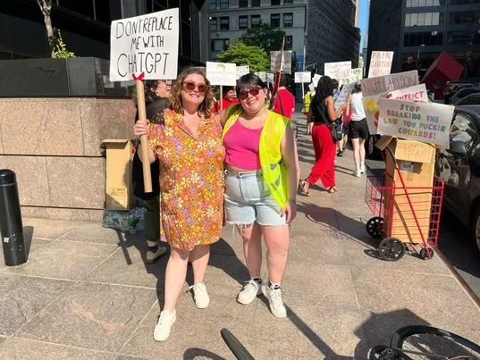 two people standing in front of a picket line. the person on the left is holding a sign that says "don't replace me with chat gpt." a sign in a box reads "stop breaking the law you fuckin cowards!"