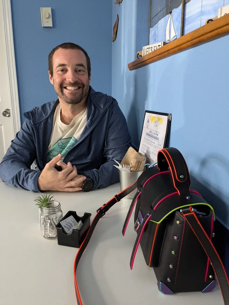 A man with a beard and short hair, smiling, sitting at a table in a blue-walled room with nautical-themed decor. On the table, there is a small potted plant and a MK black bag with neon accents.