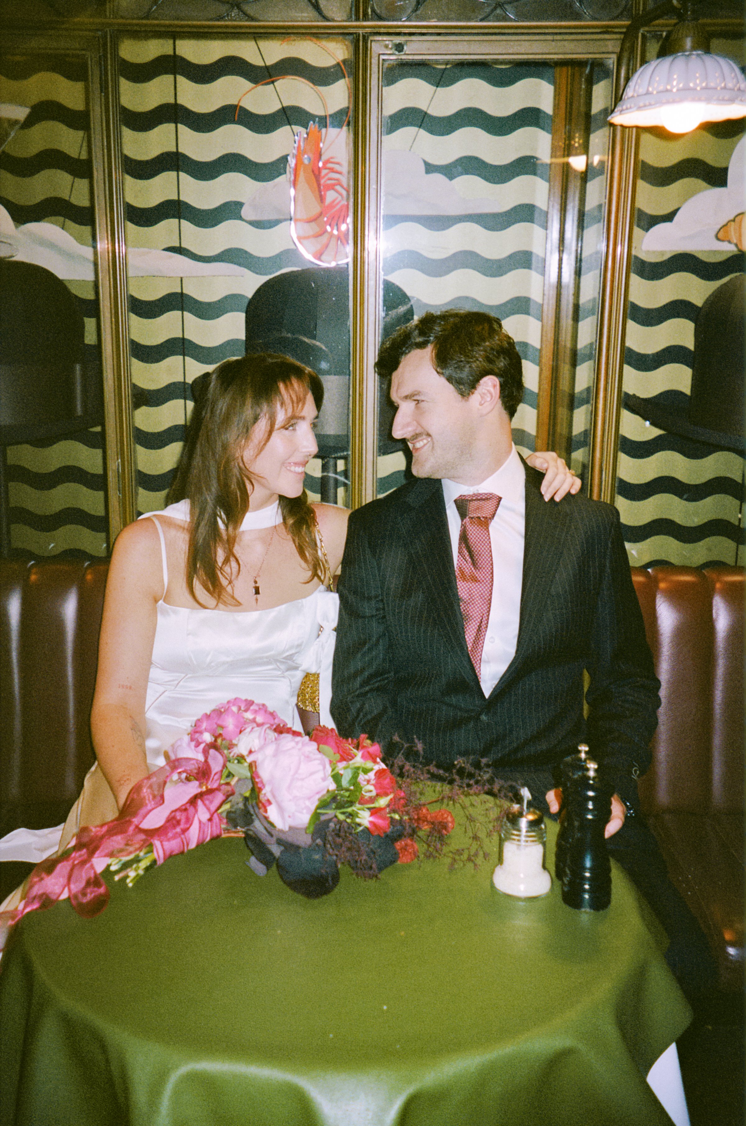 A woman and a man are sitting close together at a table, smiling and looking at each other. The woman is wearing a white dress and holding a bouquet of pink and red flowers. The man is in a dark striped suit with a pink tie. The table has a green tablecloth and is decorated with a floral arrangement, salt and pepper shakers, and a pepper grinder. The background features a wall with a wavy pattern, mirror panels, and fish and cloud illustrations.
