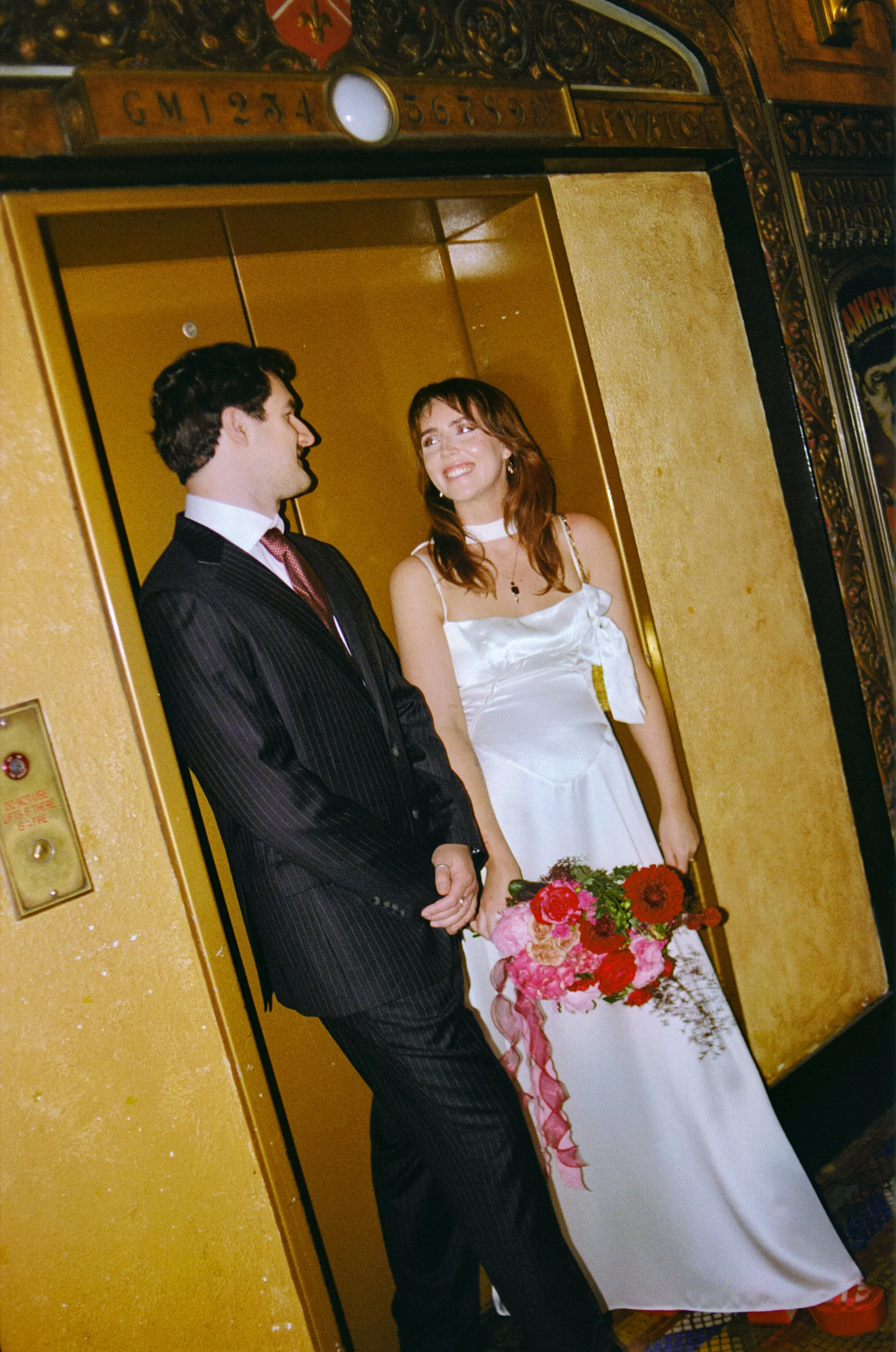 Analogue image of a couple at The QT hotel with man in a black suit and a woman in a white dress holding a bouquet of flowers standing in an elevator, smiling at each other.