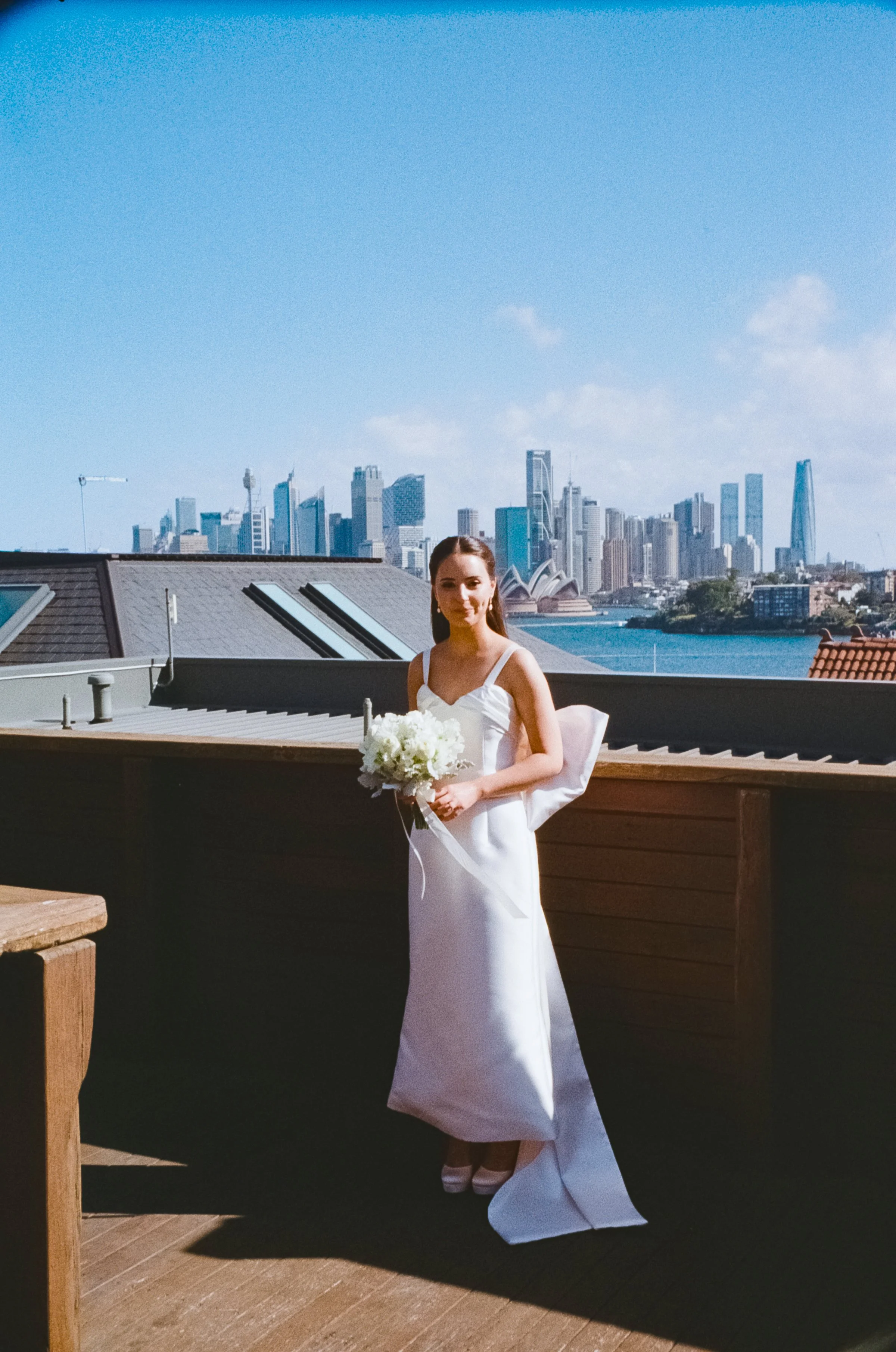 A woman in a white wedding dress holding a bouquet of white flowers on a rooftop with city skyscrapers and Sydney Opera House in the background under a blue sky.