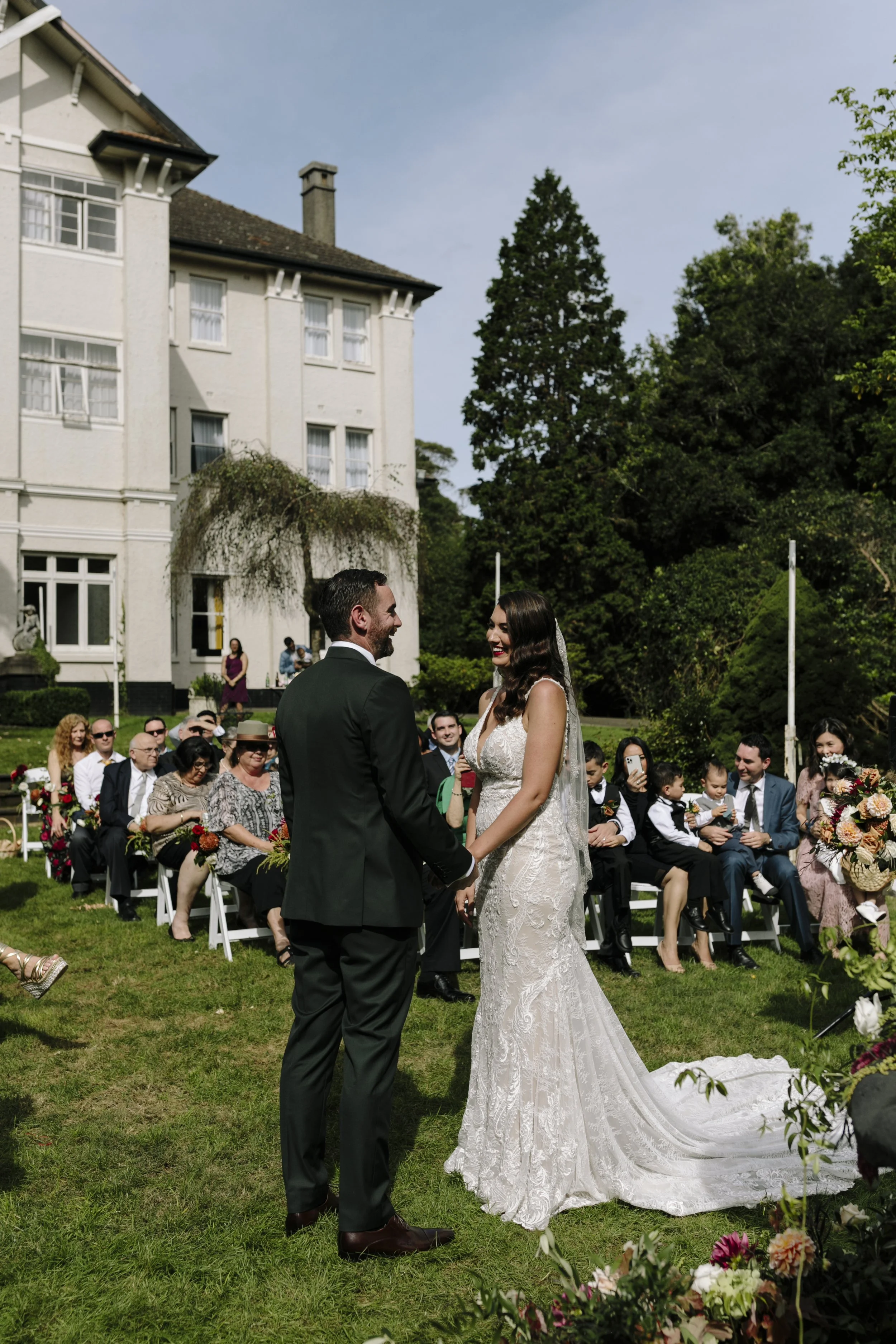 A bride and groom stand facing each other at an outdoor wedding ceremony. The bride is wearing a white lace wedding gown with a veil, and the groom is in a black suit with a white shirt. Guests are seated behind them on white chairs, and a large house with trees surrounds the ceremony.