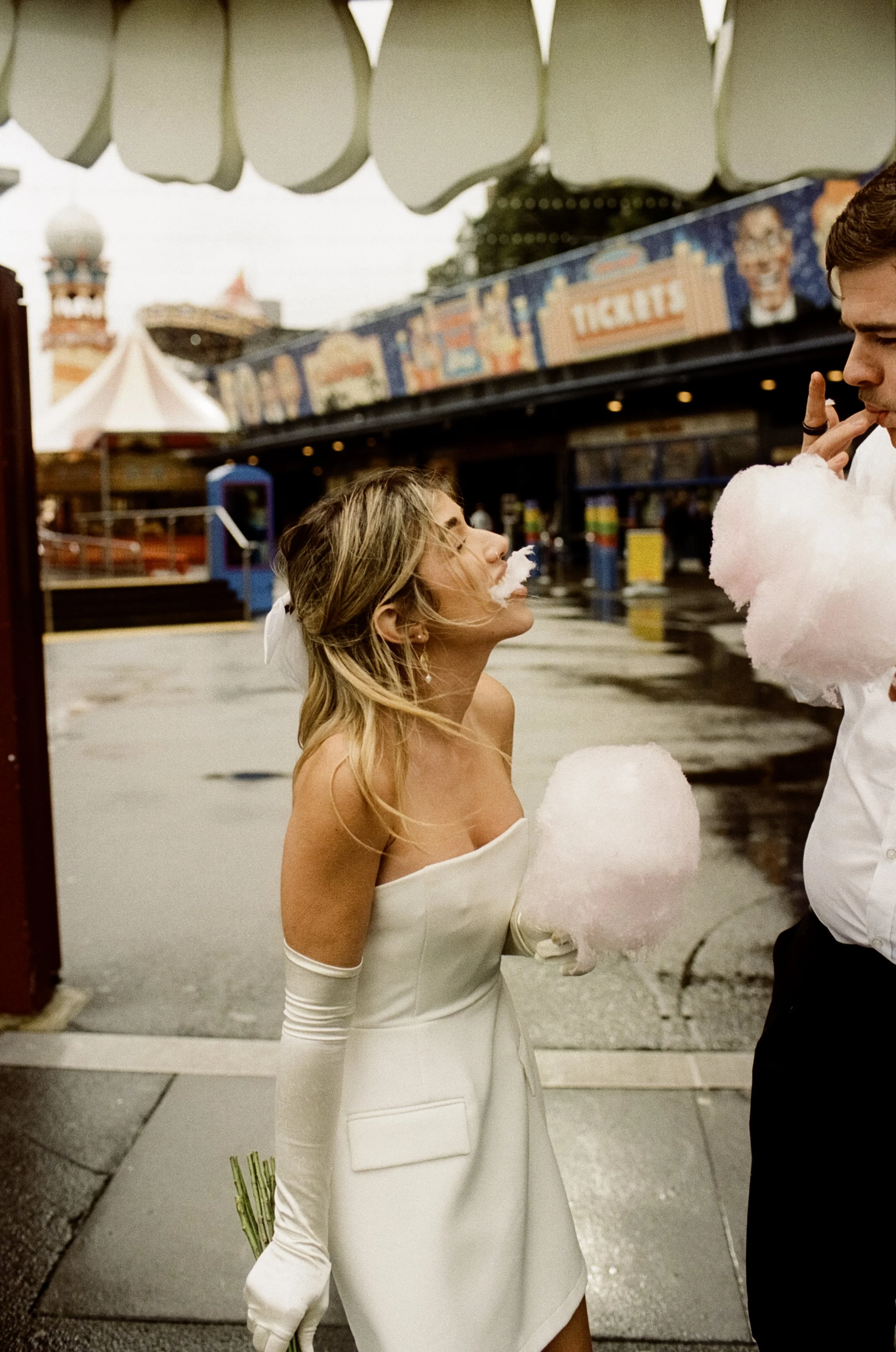 Analogue image at Luna Park Sydney with a bride in a white off-the-shoulder wedding gown holding a bouquet of flowers, with blonde hair, sharing cotton candy with a man at a carnival or amusement park.