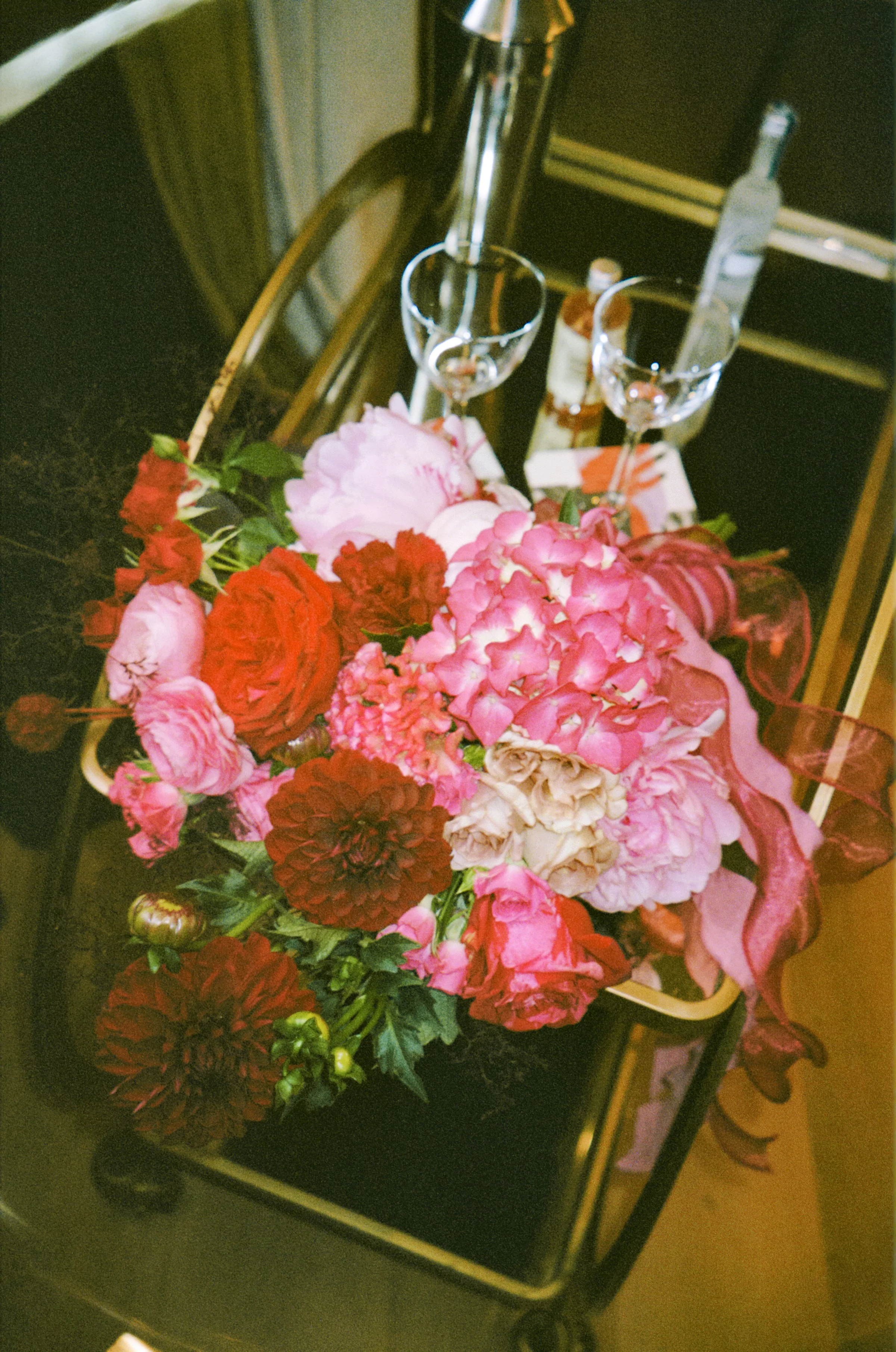 A black cart with gold trim containing a large bouquet of red, pink, and cream flowers, and two empty wine glasses, some bottles, and a white tabletop with napkins and utensils.