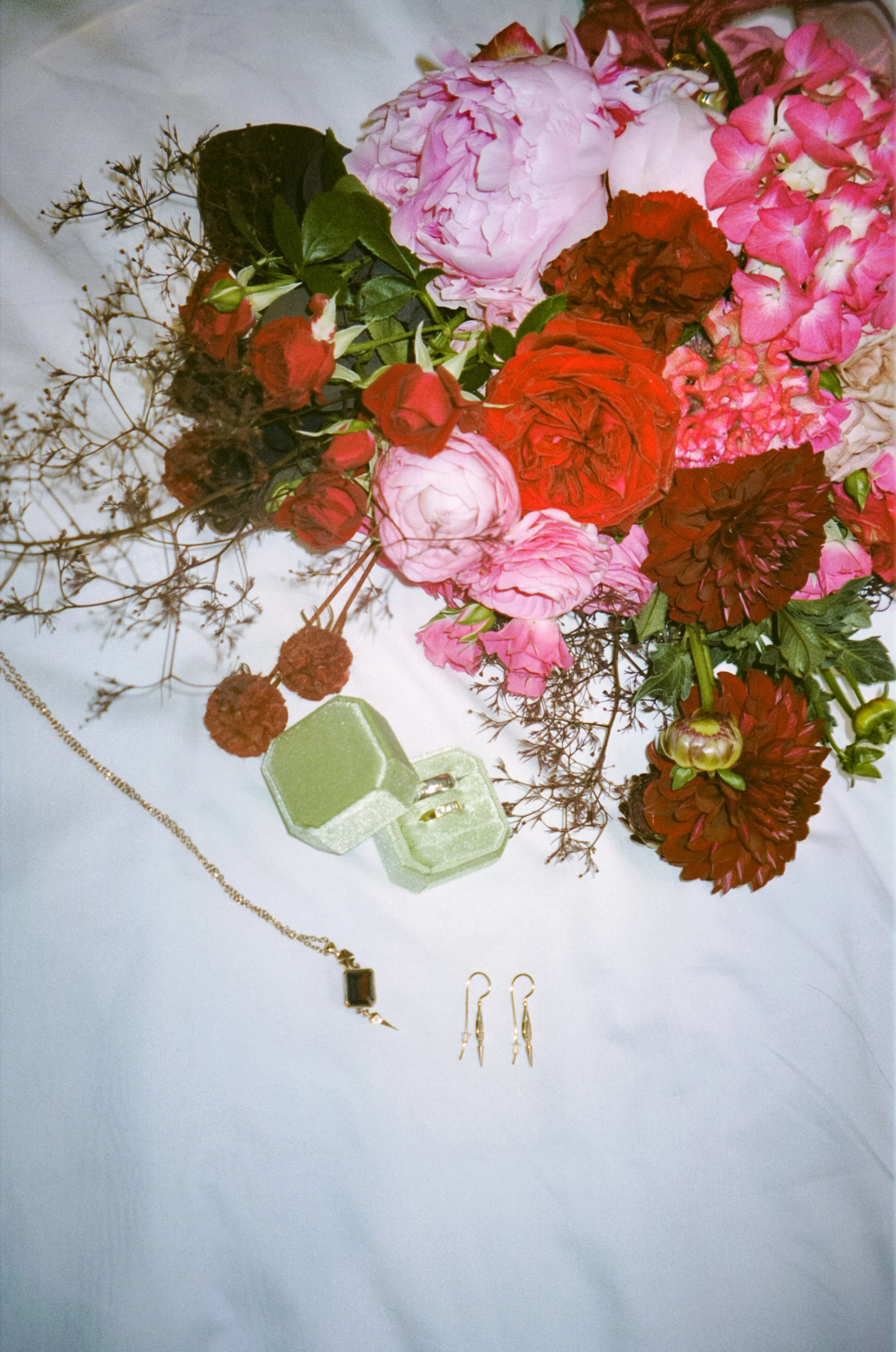 A bouquet of pink and red flowers with green leaves, a necklace with a dark stone pendant, and a pair of gold earrings are arranged on a white surface. A small green box is also visible.