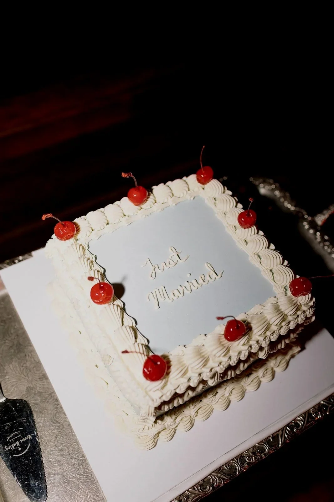 Square birthday cake with white frosting, decorated with whipped cream border and red cherries on top, with the message 'Just Married' written on the top surface.