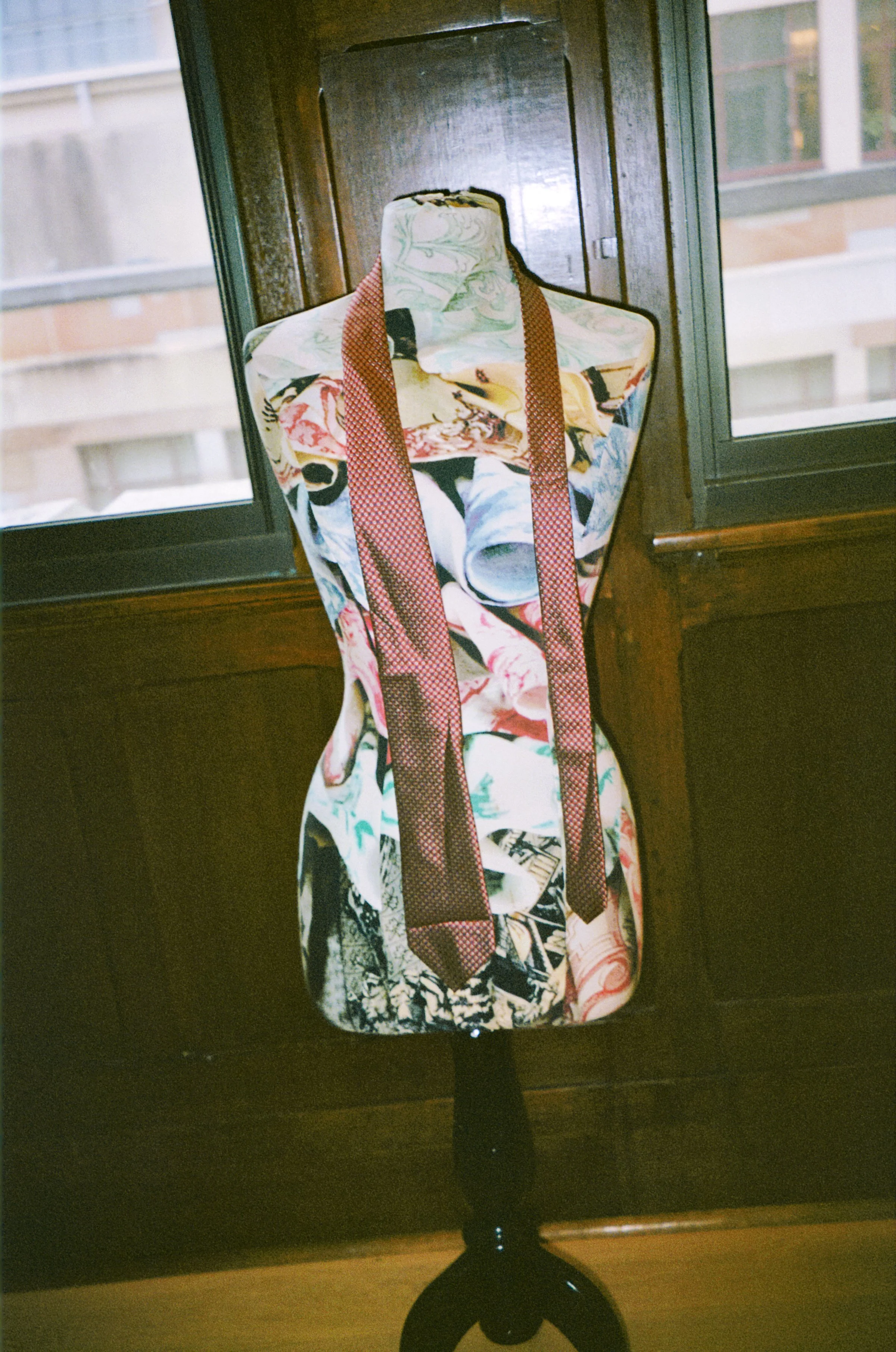 A decorative mannequin bust on a black stand, draped with a colorful fabric featuring various artistic patterns, positioned in front of a window with wooden trim.