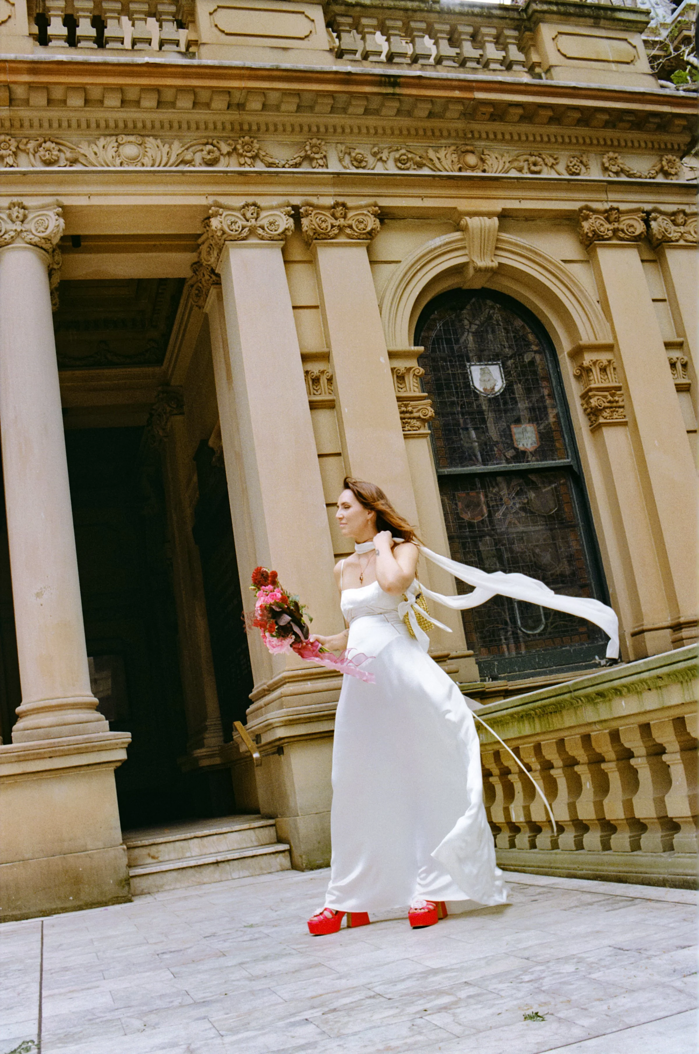 analogue image of a woman in a white dress and red platform shoes holding a bouquet of pink and red flowers stands outside a historic building Town Hall Sydney with ornate columns and stained glass windows.
