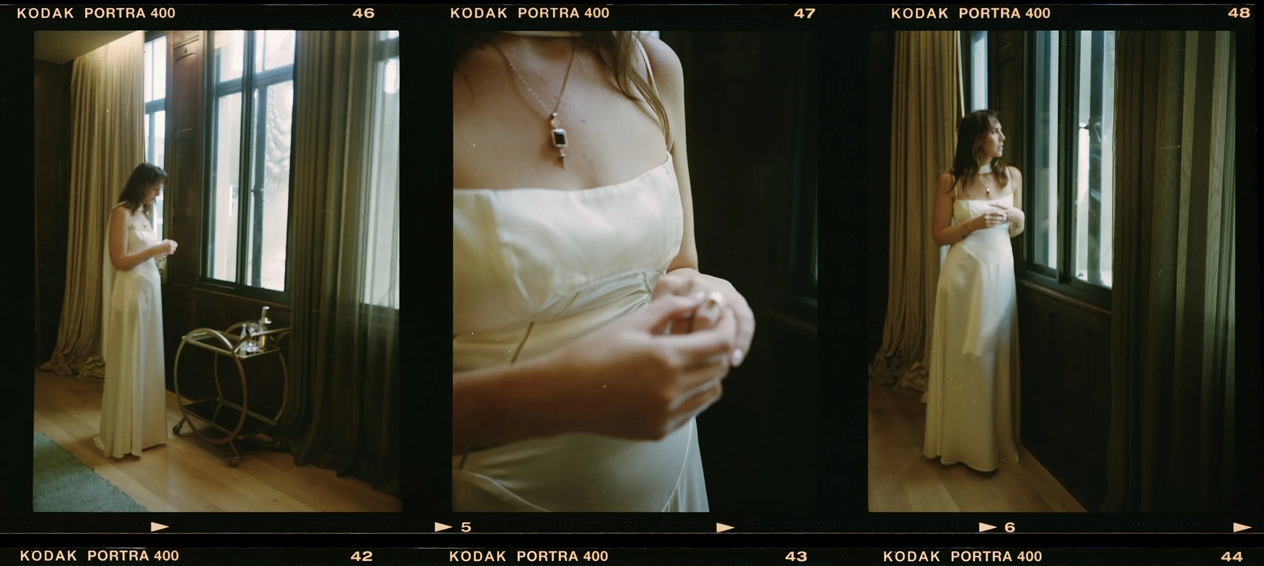 A woman in a white satin gown standing by a large window, looking outside, with a drink on a small table beside her, during daytime.