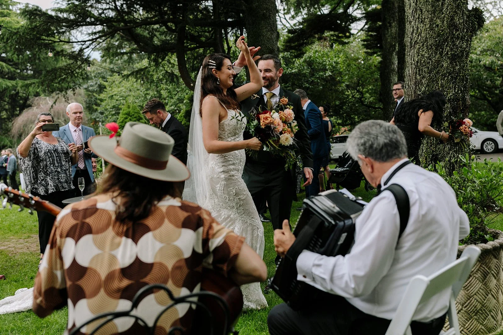 A wedding celebration outdoors with the bride and groom dancing, surrounded by guests, trees, and greenery.