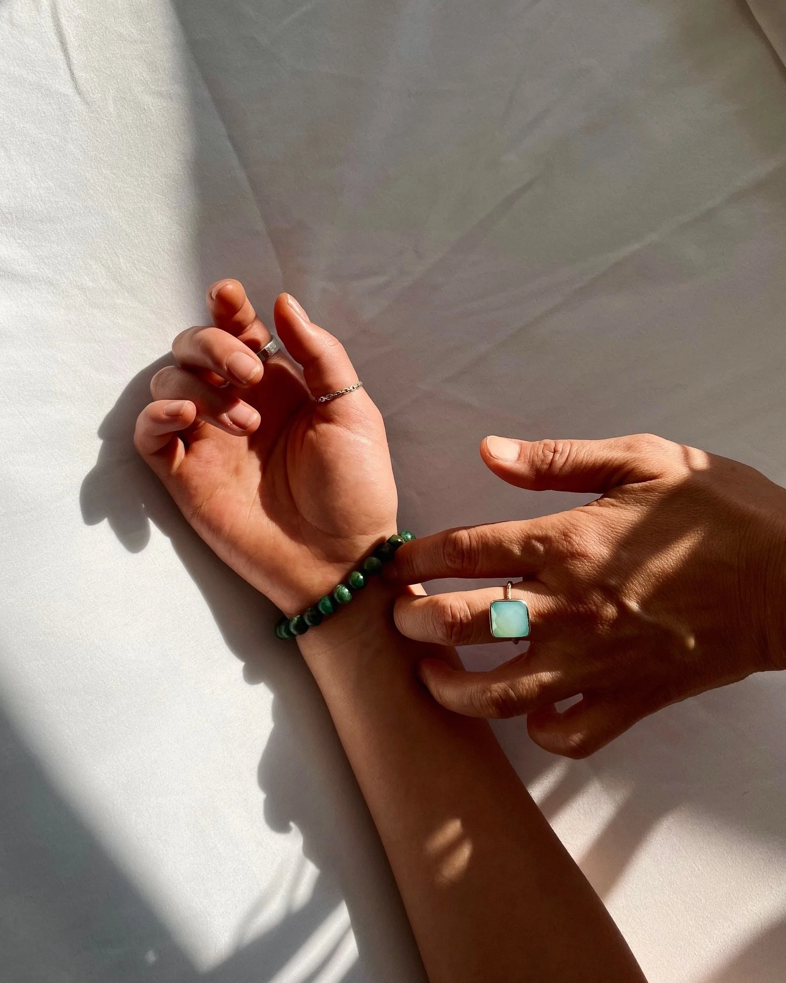 Close-up of two hands, one resting on a white surface with shadows, the other gently taking the pulse of the first hand. The hands are adorned with rings and a beaded bracelet, with sunlight casting shadows.