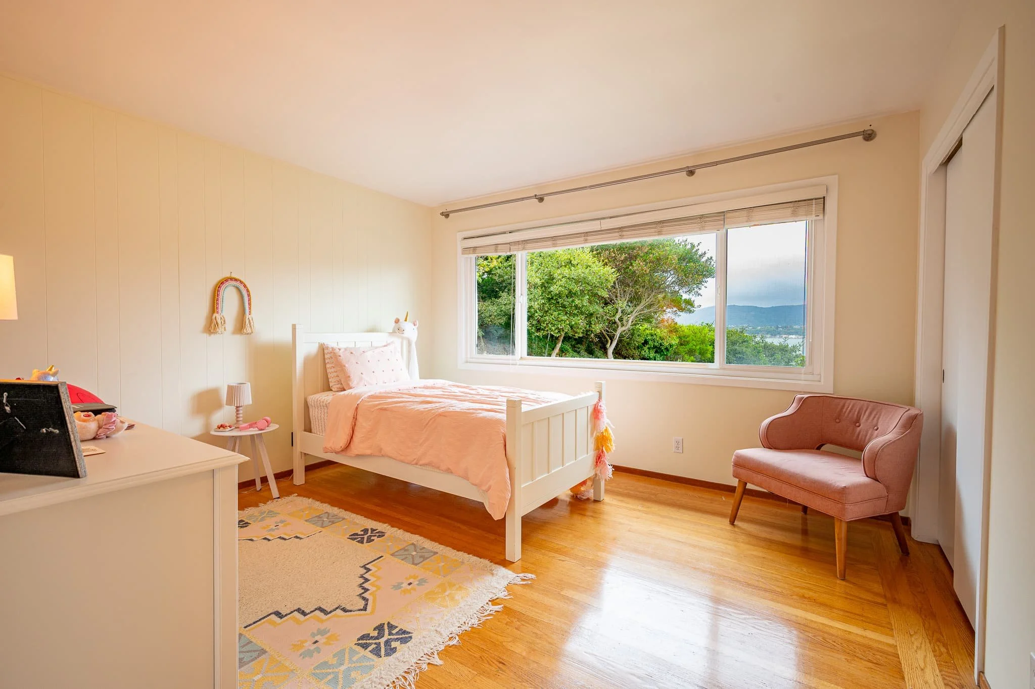 A bedroom with a large window showing green trees outside, a white bed with pink bedding, a pink armchair, and a multi-colored geometric rug.